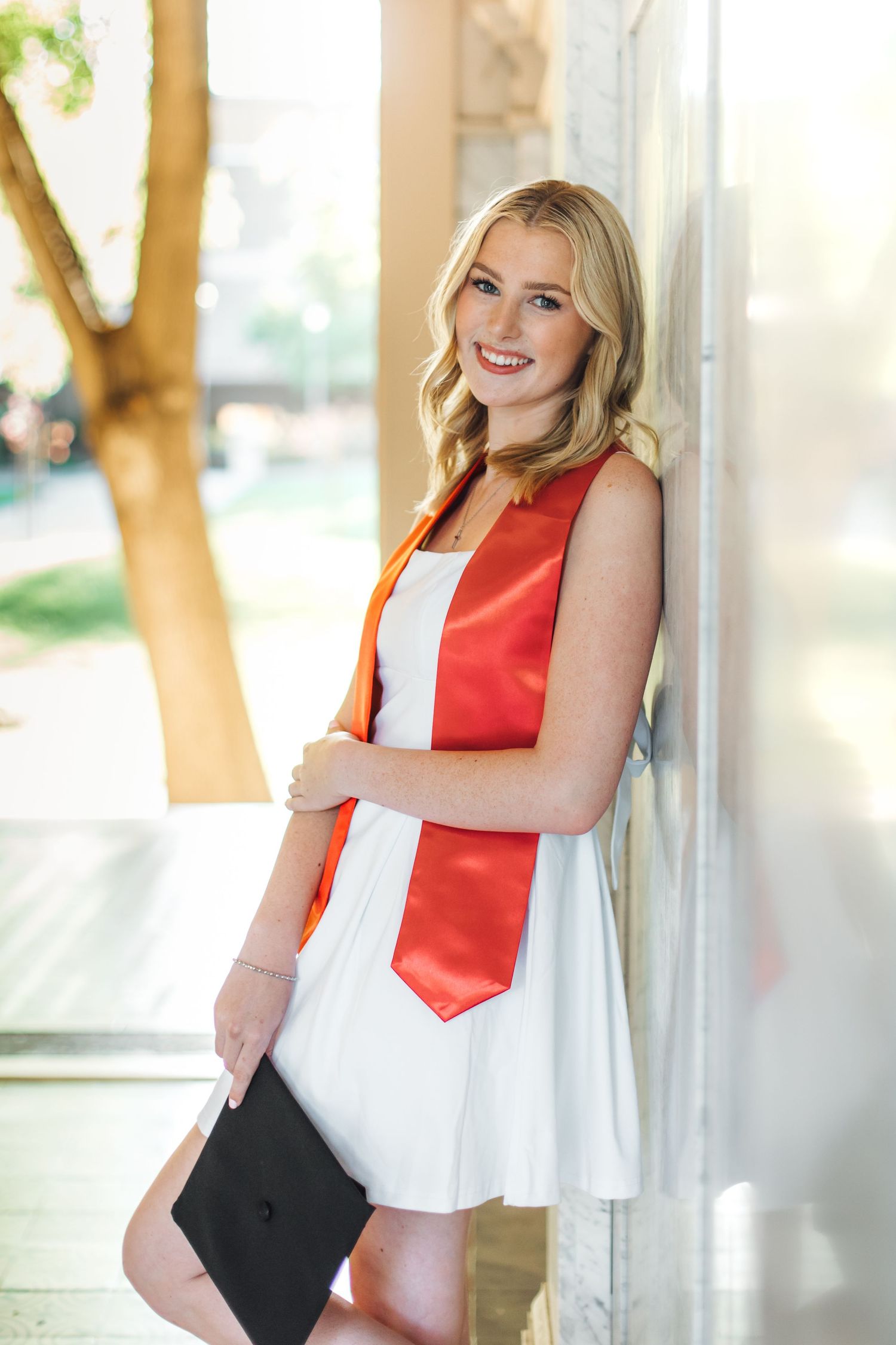 Smiling blonde graduate in white dress and red stole leans against wall holding graduation cap.
