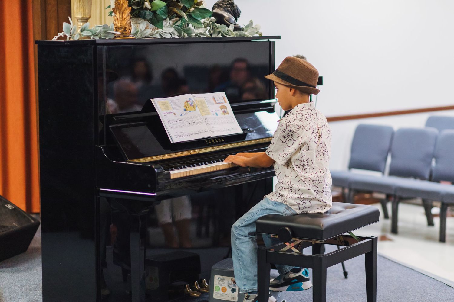A boy in a hat plays an upright piano at a recital.