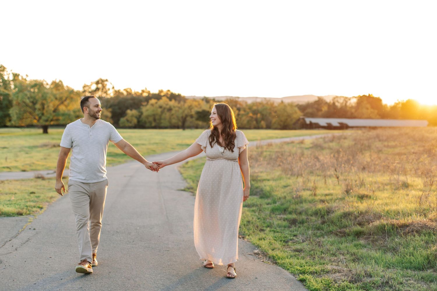 A couple holds hands walking down a sunlit path surrounded by golden fields at sunset.