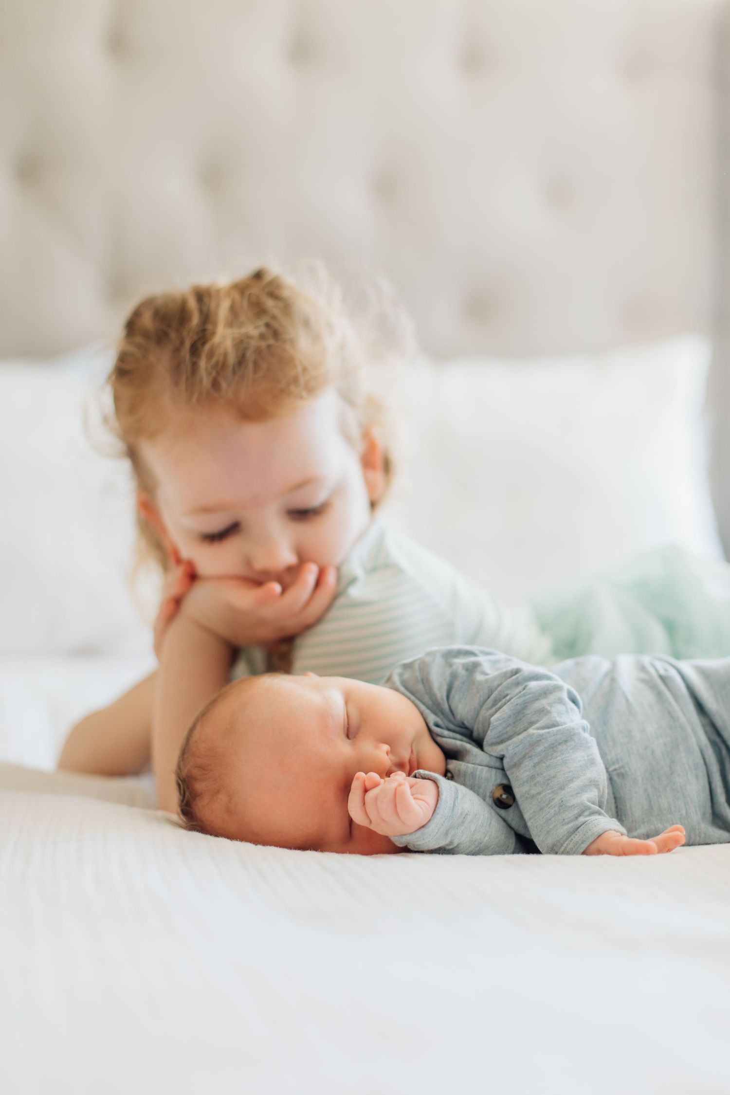 Curious toddler rests chin on hands watching sleeping newborn baby with a pacifier on a white bed.