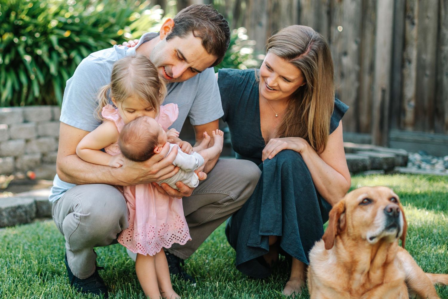 Young family with toddler daughter, newborn baby, and golden retriever dog sitting on green grass.