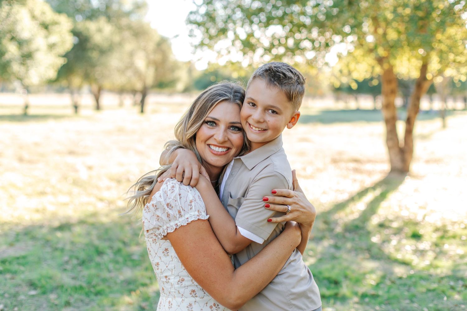 A smiling woman in a lace dress hugs a young boy outdoors in a sunlit park setting.