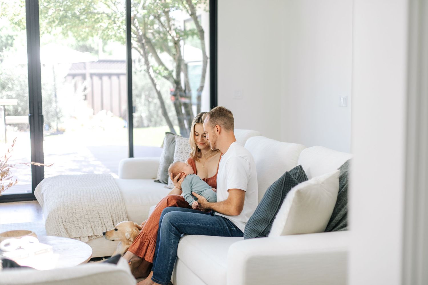 Young couple sitting closely on a white couch in a bright living room, bonding with their newborn.
