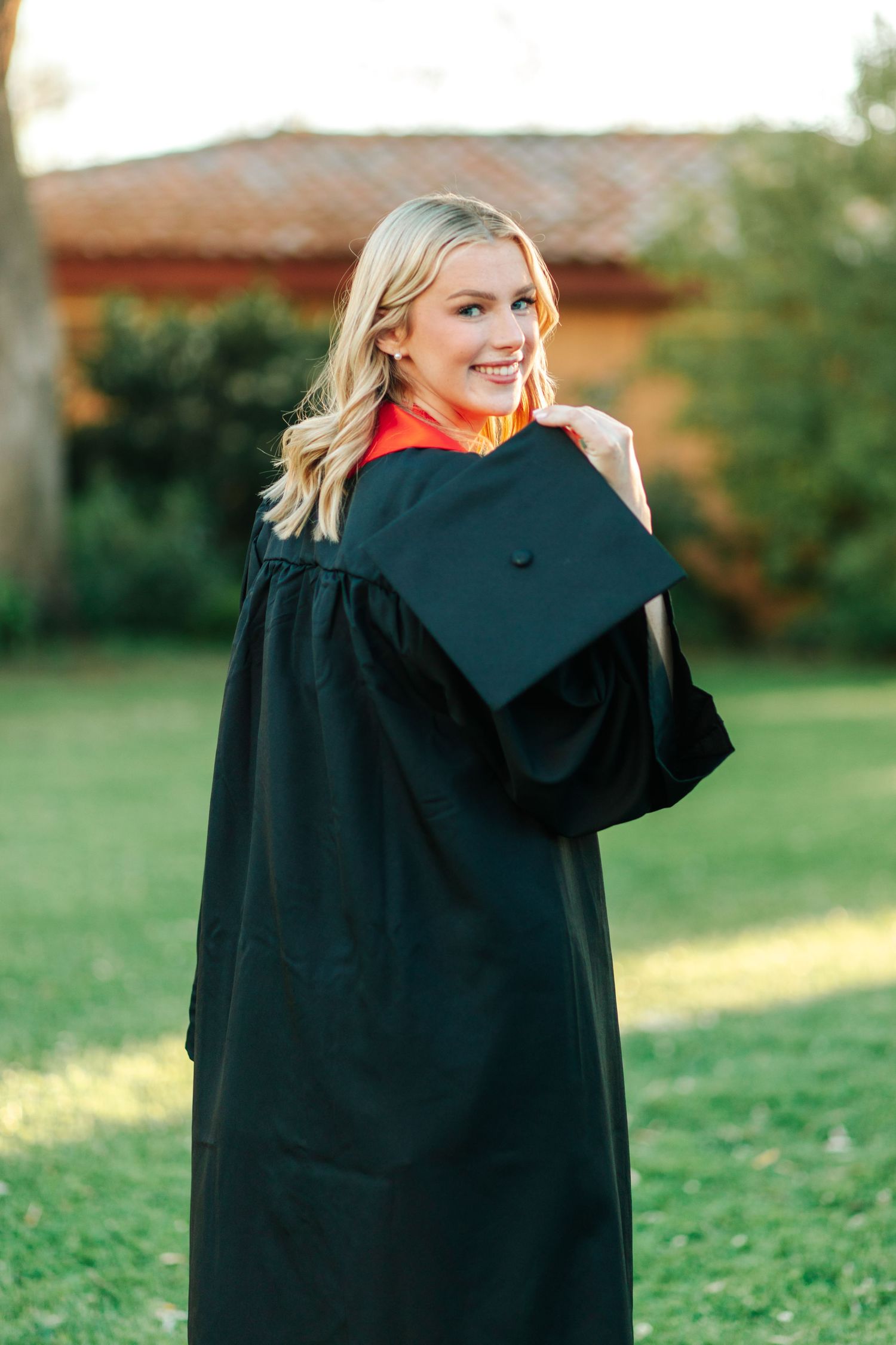 Blonde woman in black graduation gown and cap smiling over shoulder on green campus lawn.