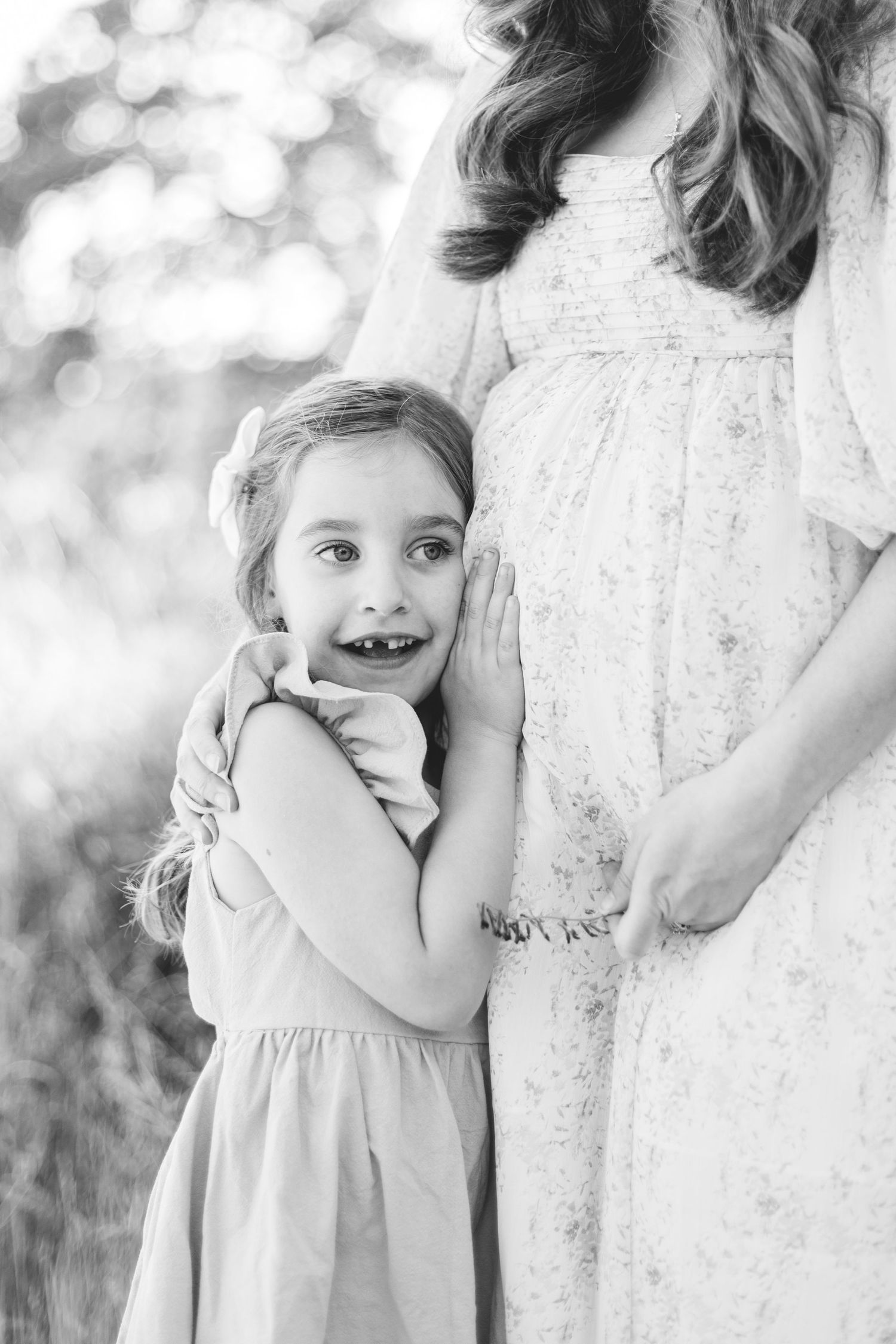 Black and white photo of a young girl hugging her pregnant mothers belly outdoors.