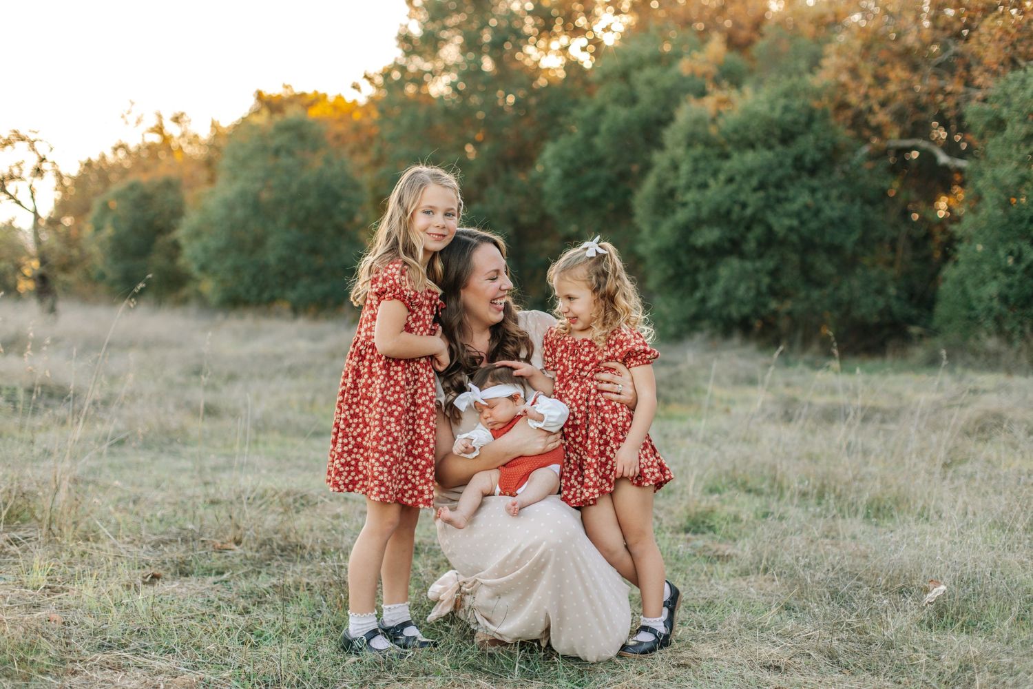 A mother kneels in a field holding a newborn while two daughters in red dresses embrace her warmly.