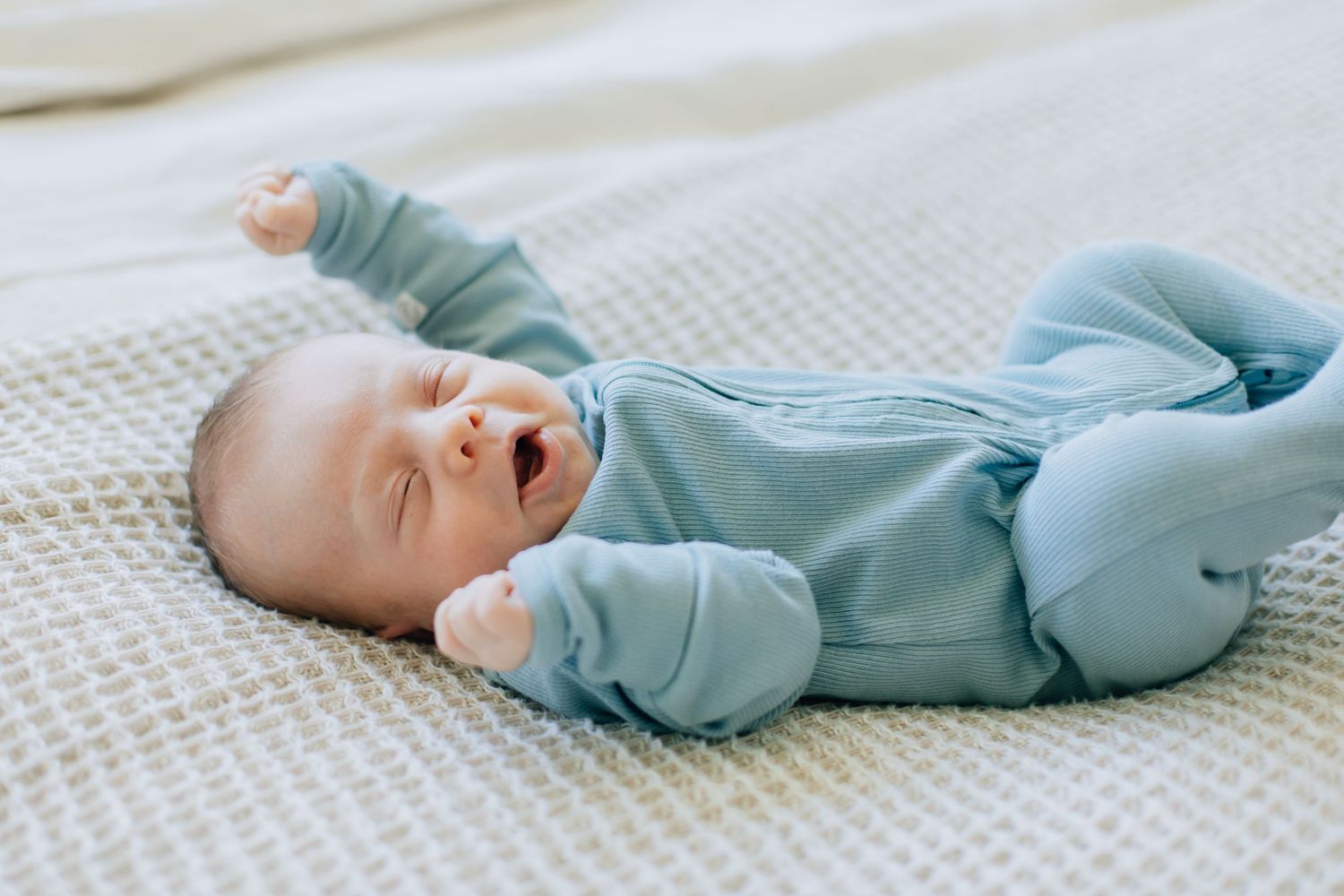 Peaceful newborn baby in blue sleepsuit lying on white knit blanket with arms stretched wide.