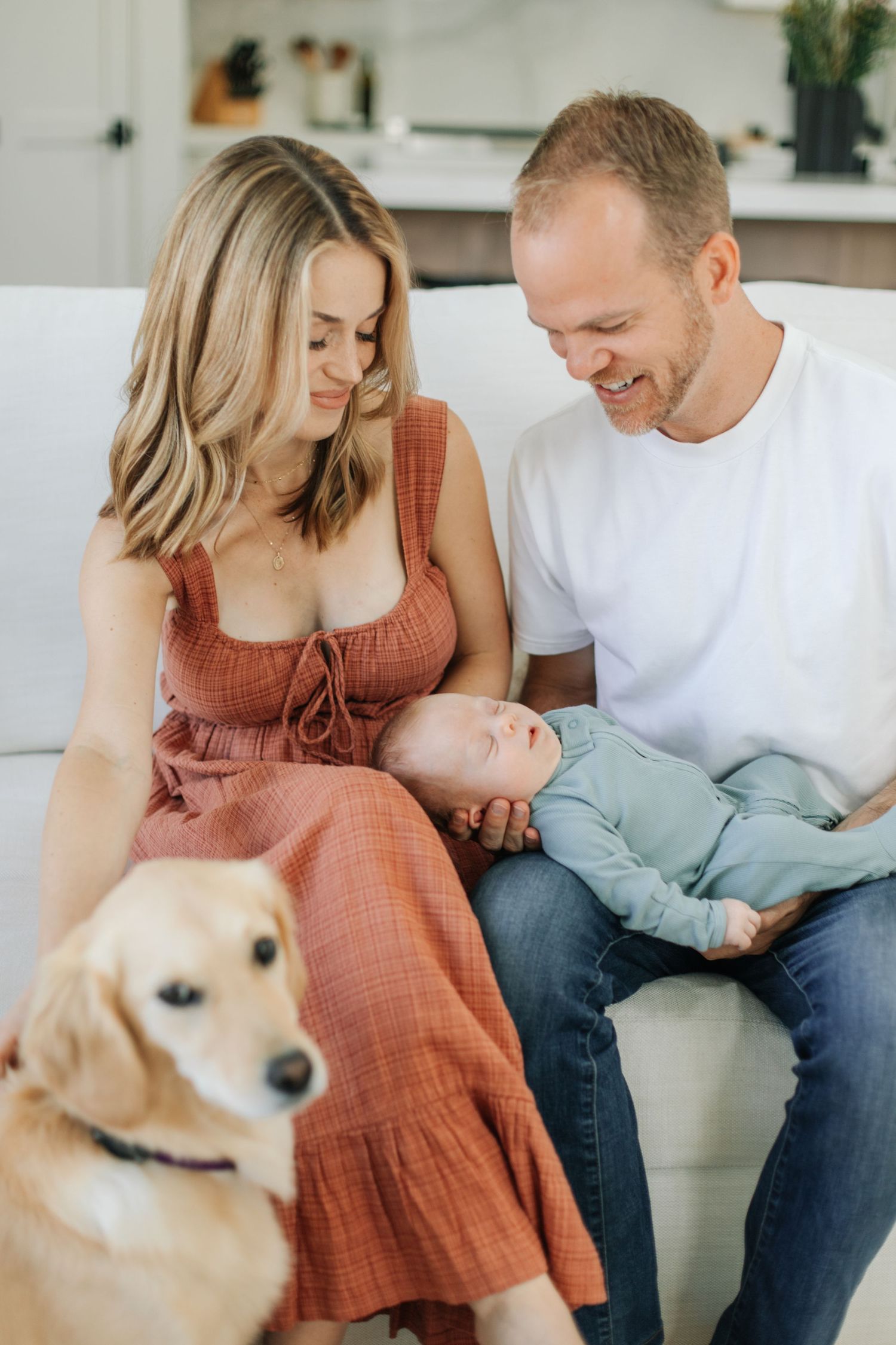 A smiling family with a newborn baby and golden retriever dog sitting on a white couch at home.
