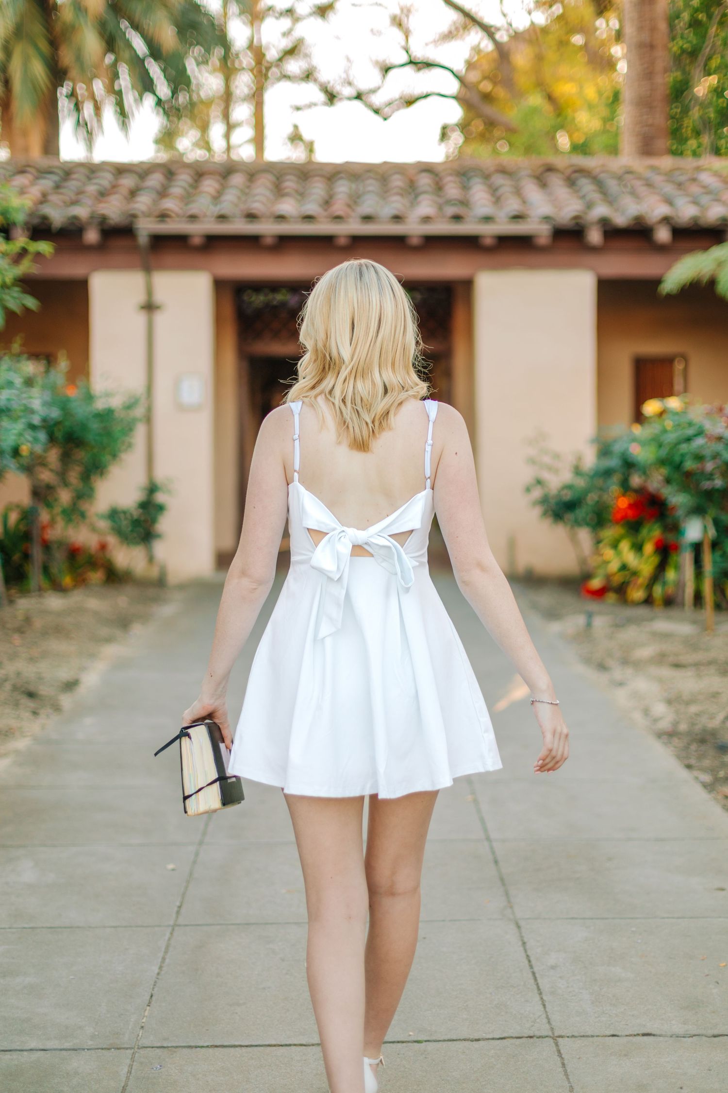 Back view of blonde graduate in white bow-back dress walking along garden pathway at sunset.