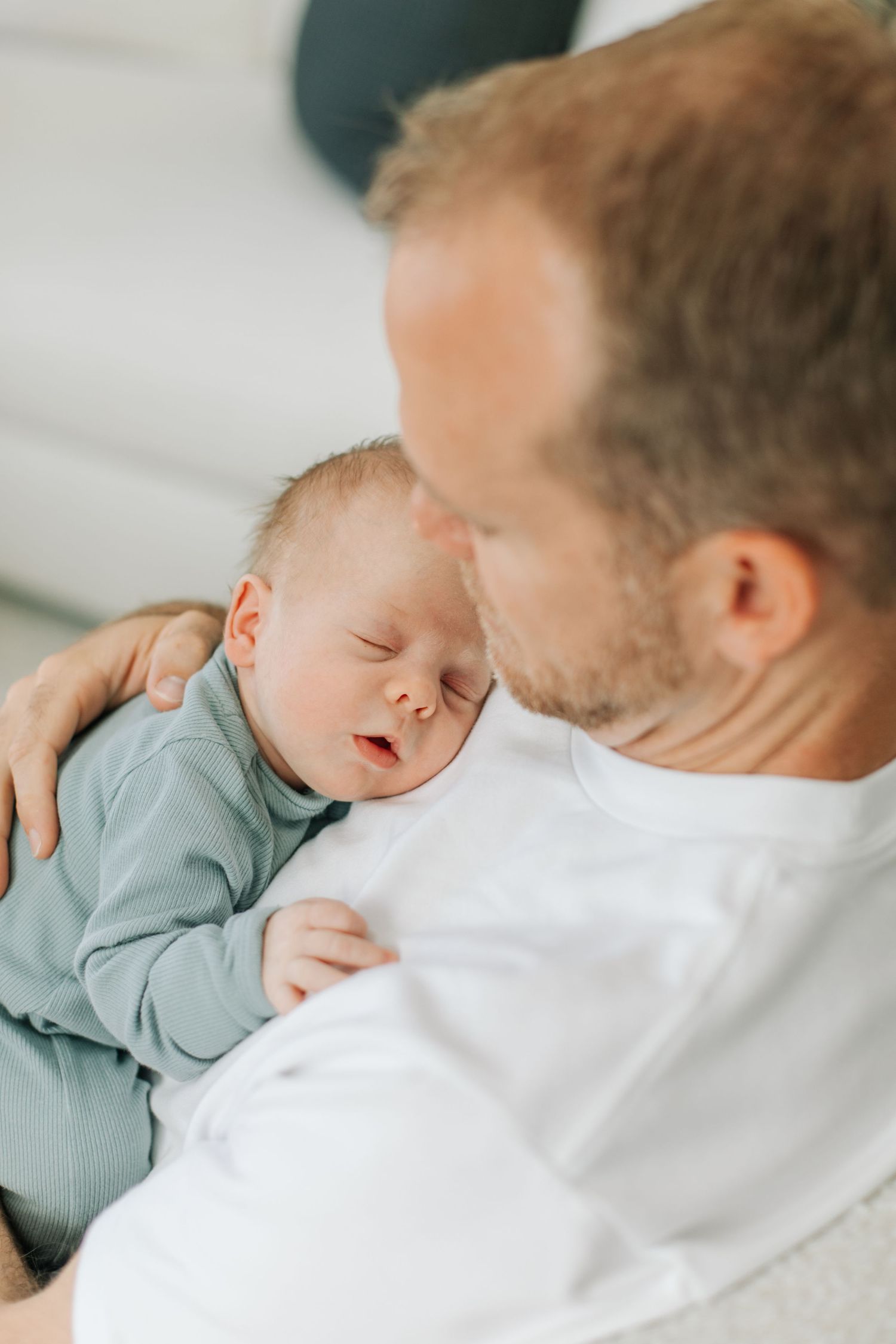 Father tenderly holding sleeping newborn baby dressed in sage green outfit close to his chest.