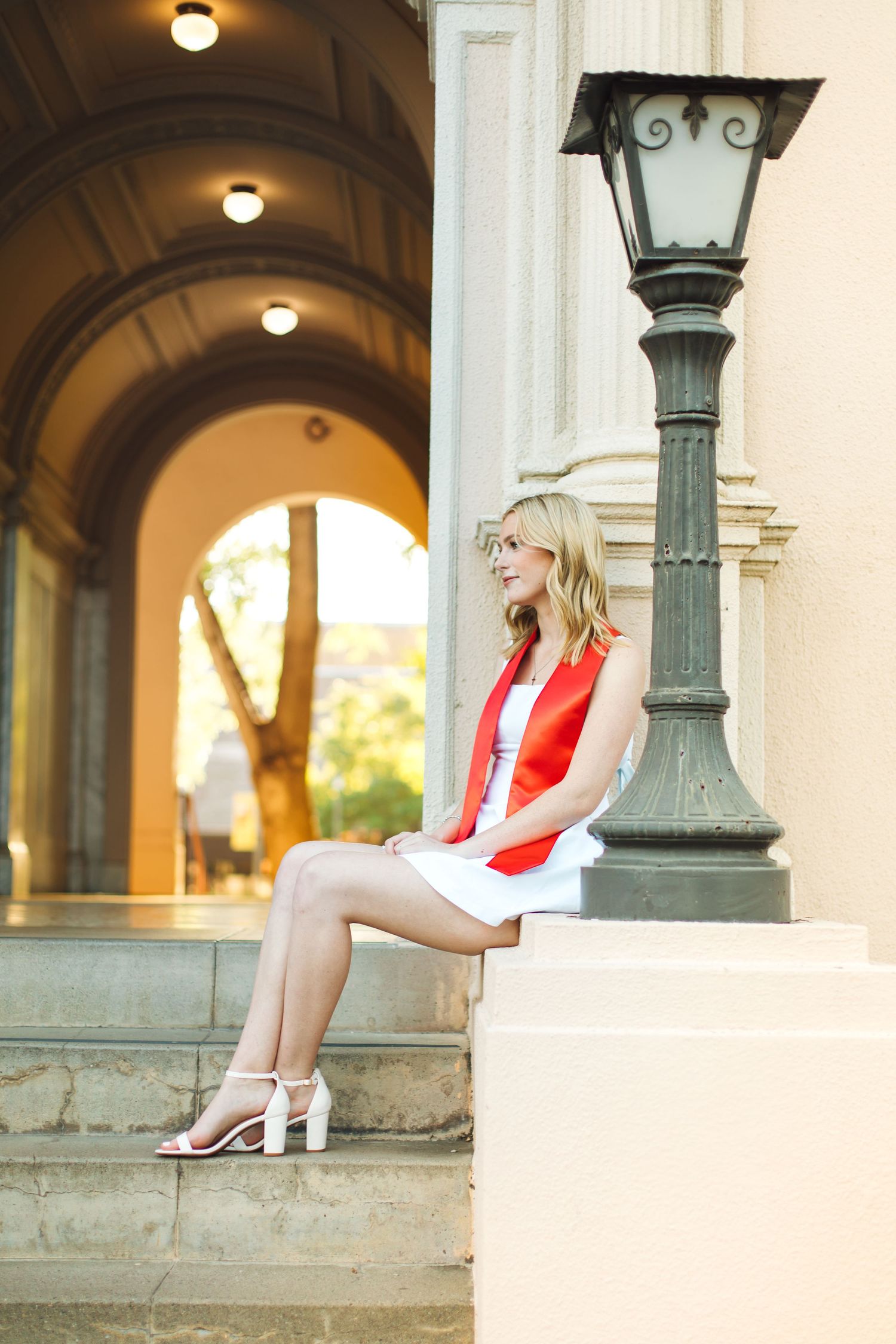Blonde graduate in white dress and red stole sits on steps beside vintage lamp post at dusk.