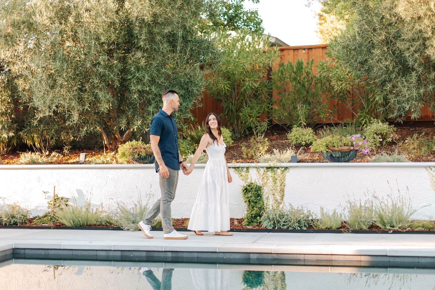 A couple holds hands and smiles while walking beside a serene backyard pool surrounded by greenery.