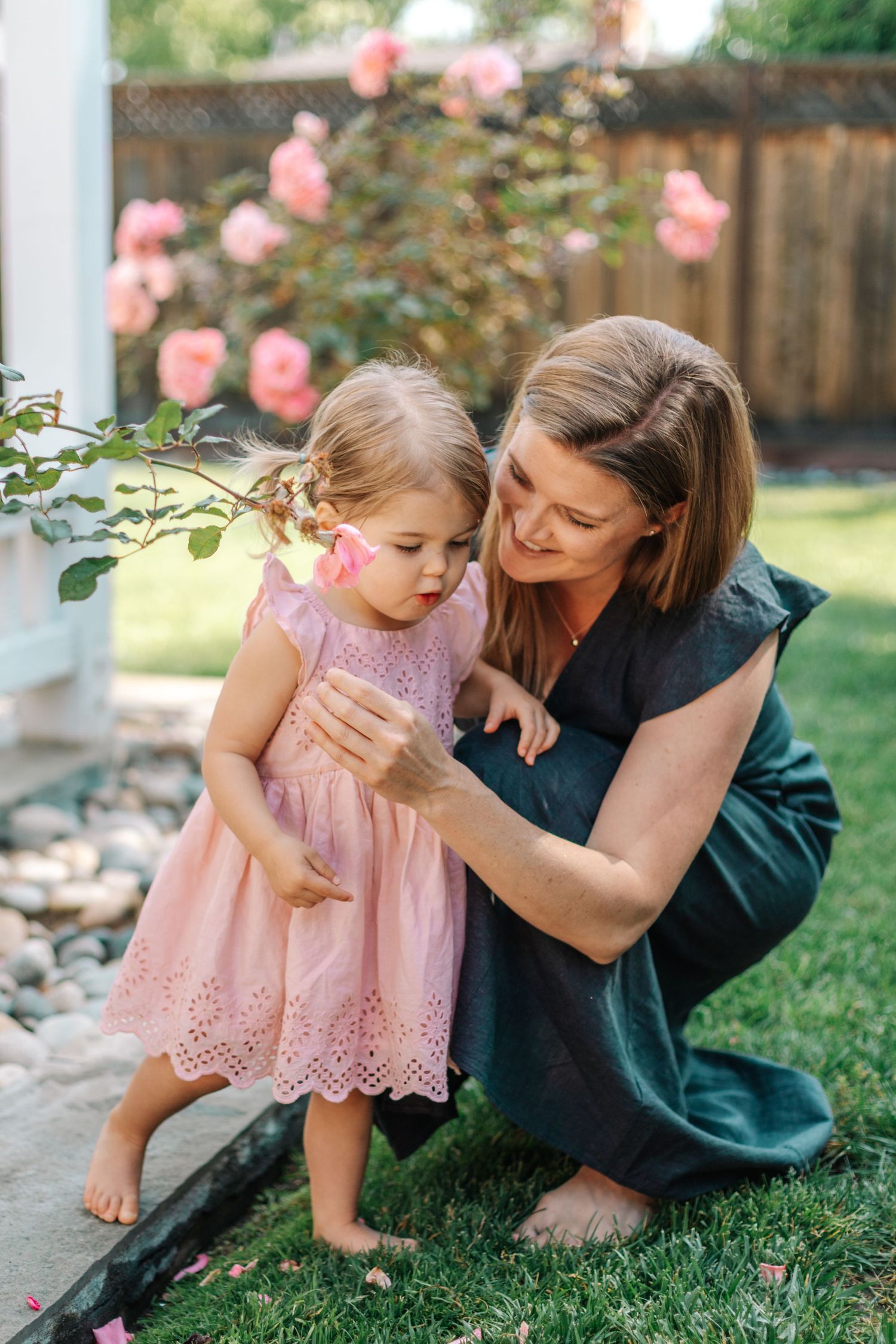 A mother crouches with her toddler daughter near pink roses in a garden.