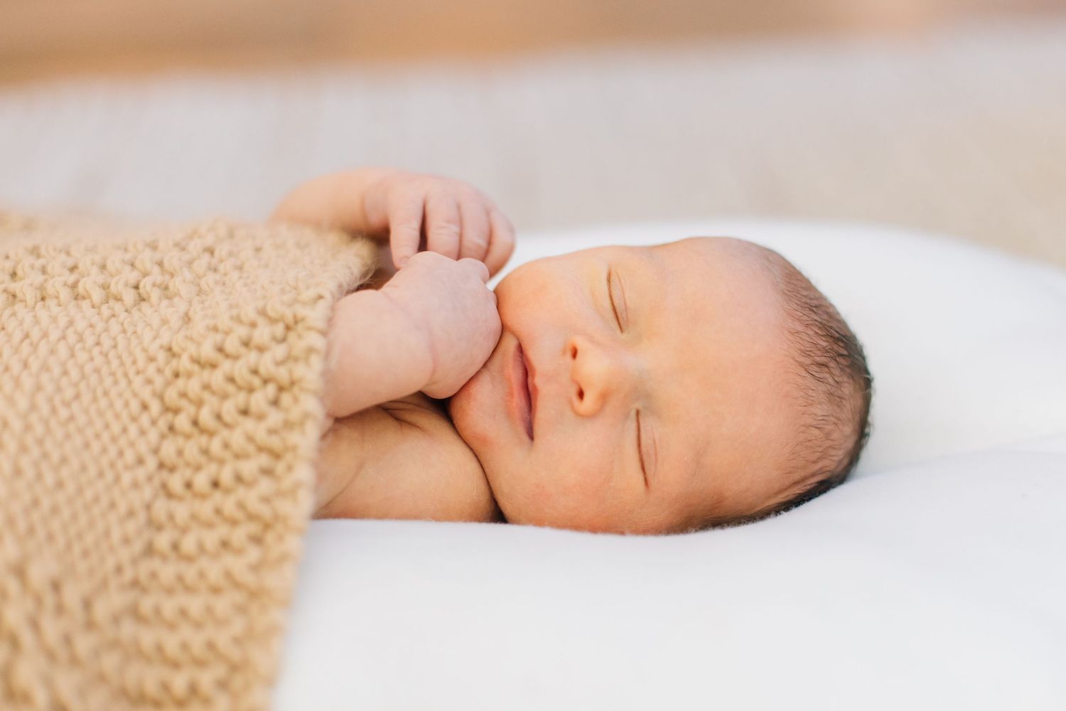 Sleeping newborn baby wrapped in tan knit blanket rests on white surface with tiny fist raised.