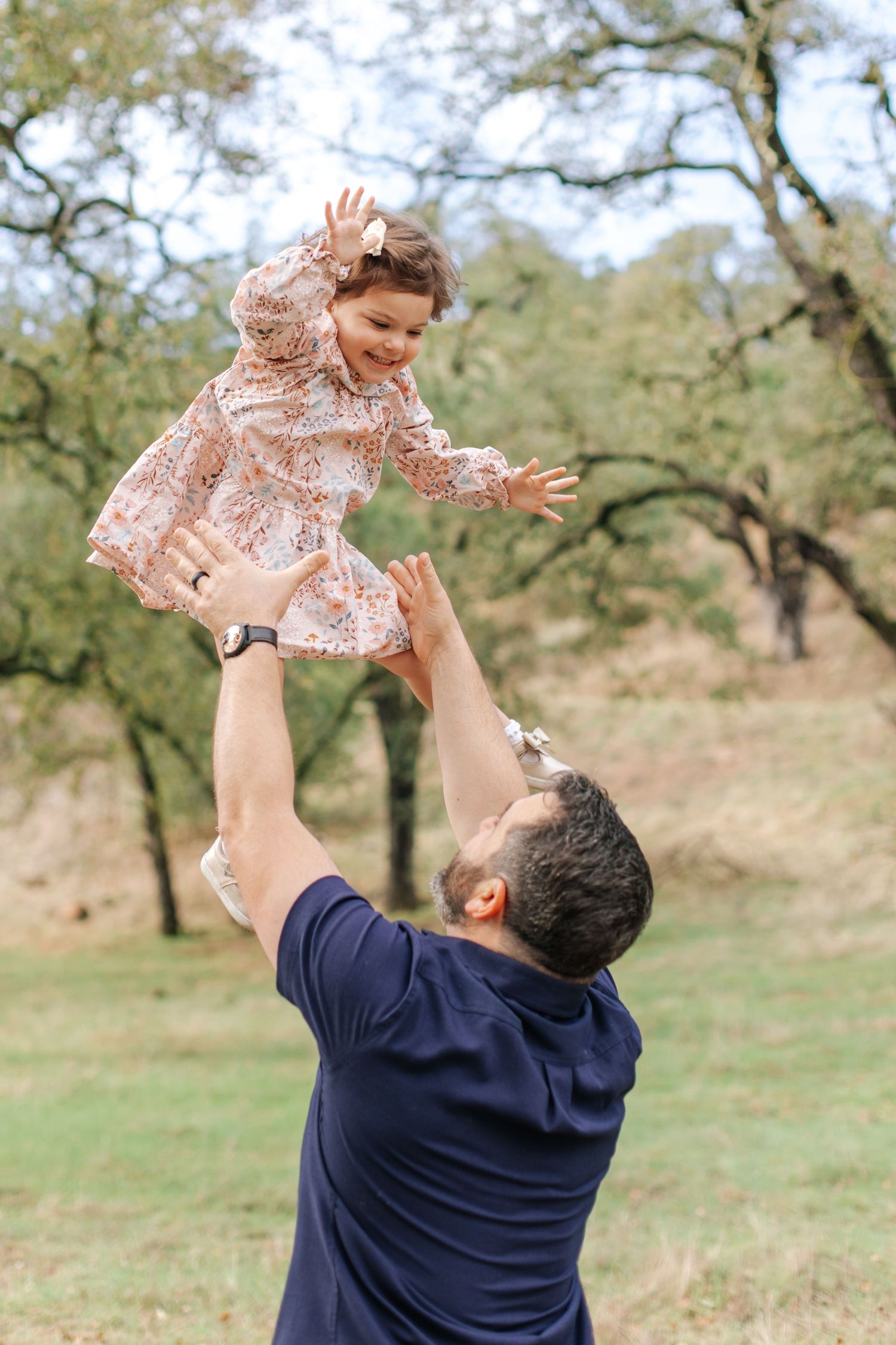 A father joyfully tosses his smiling toddler daughter in the air outdoors among trees.