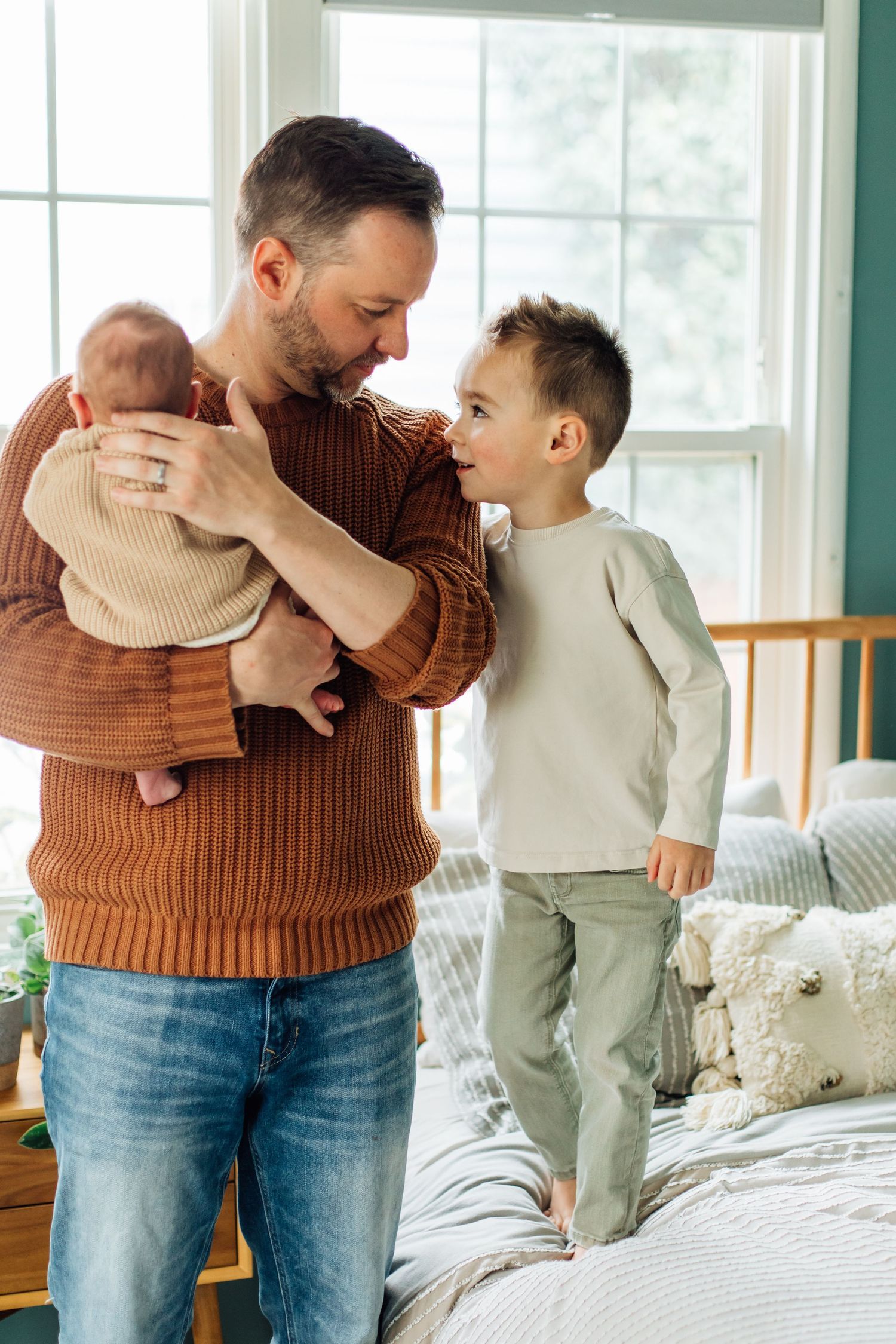 A father in a brown knit sweater holds his newborn while his young son stands on the bed.