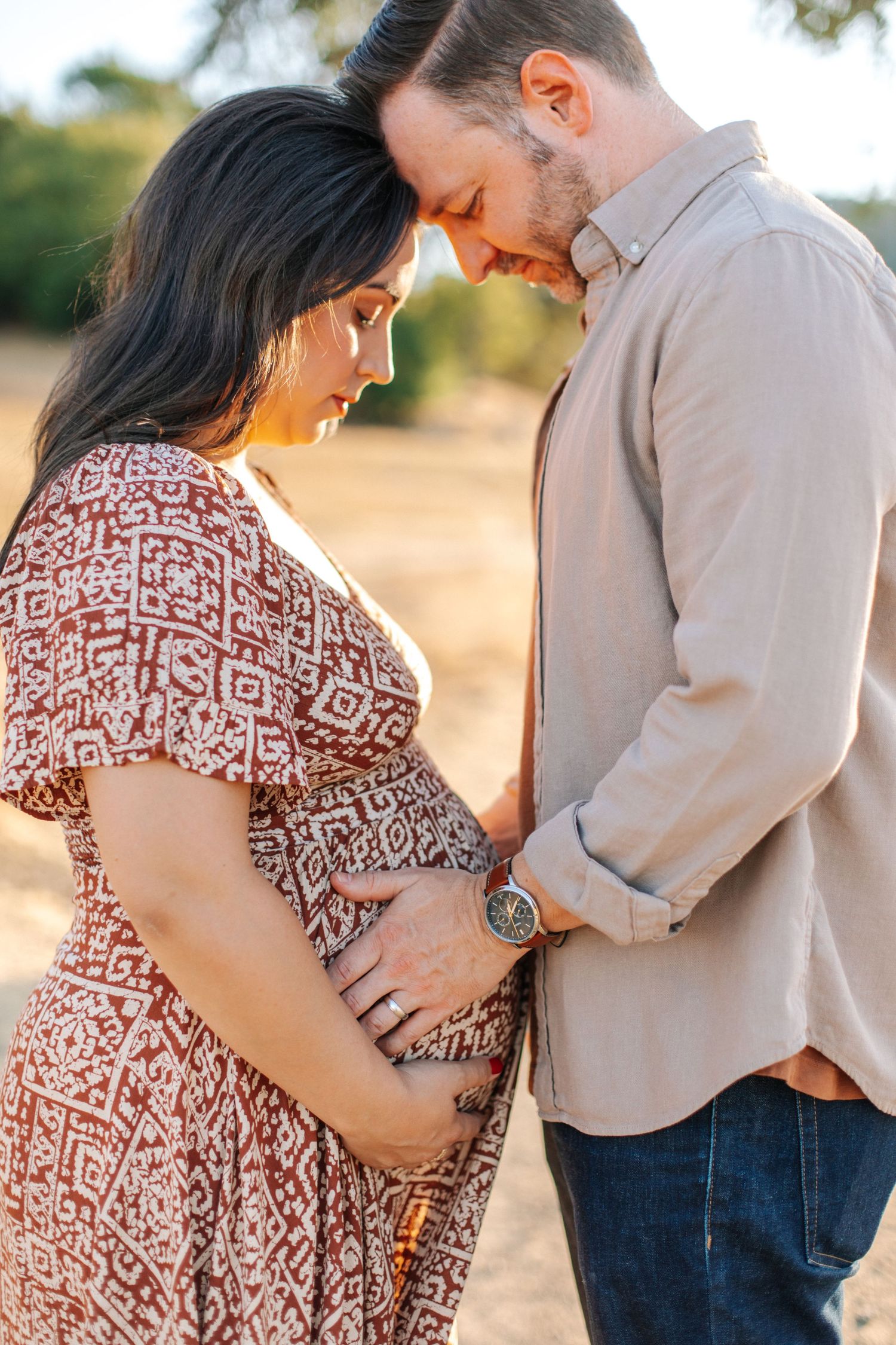 Couple tenderly touching pregnant belly together outdoors in warm golden hour light.