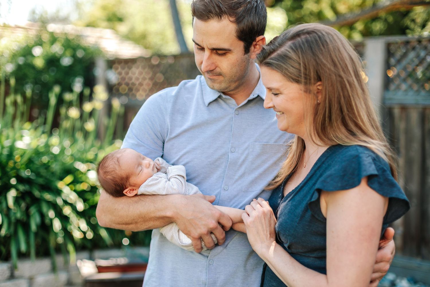 Young couple standing outdoors in lush garden smiling and looking down at their newborn baby.