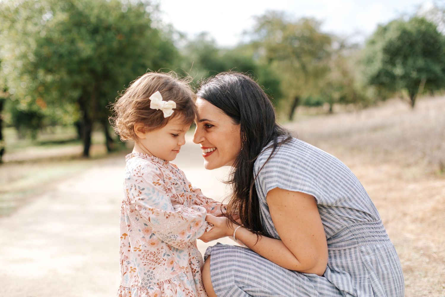 A dark-haired mother smiles face-to-face with her toddler daughter on a tree-lined dirt path.