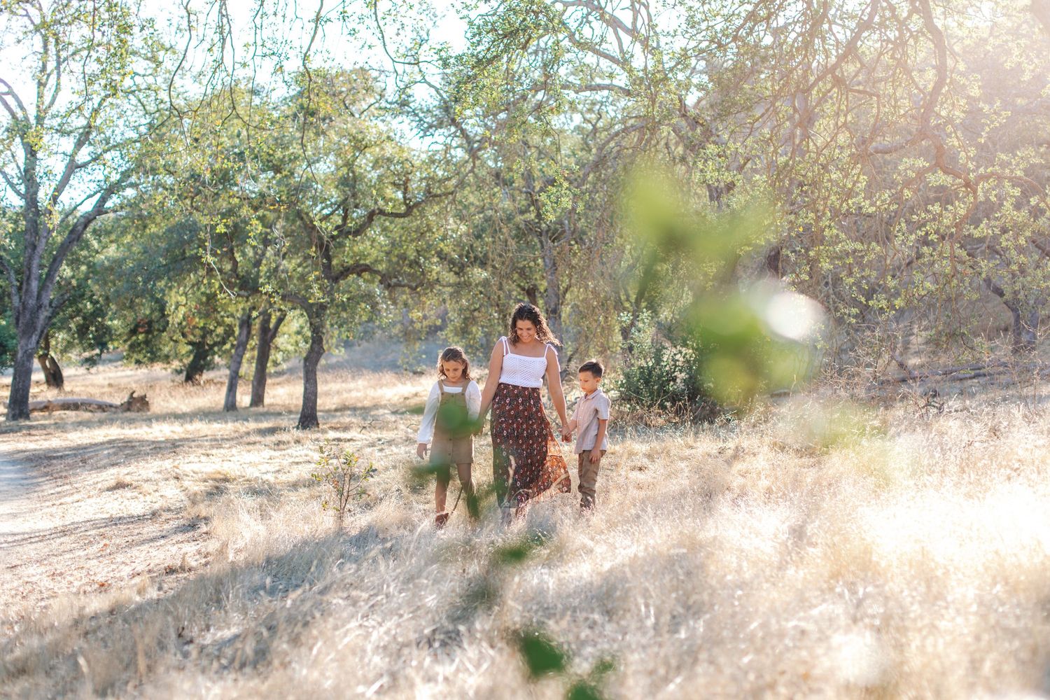 A mother walks with her children through a sunny orchard with dry grass and leafy trees in the background.