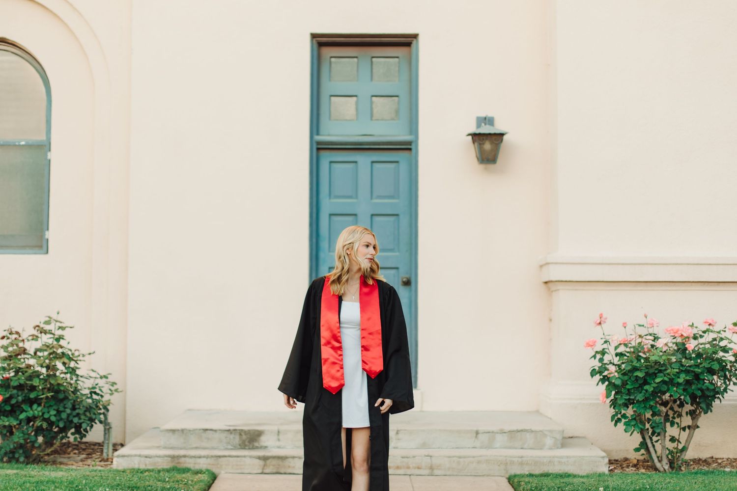 Blonde woman in black graduation gown with red stole standing before a teal door on a cream building.