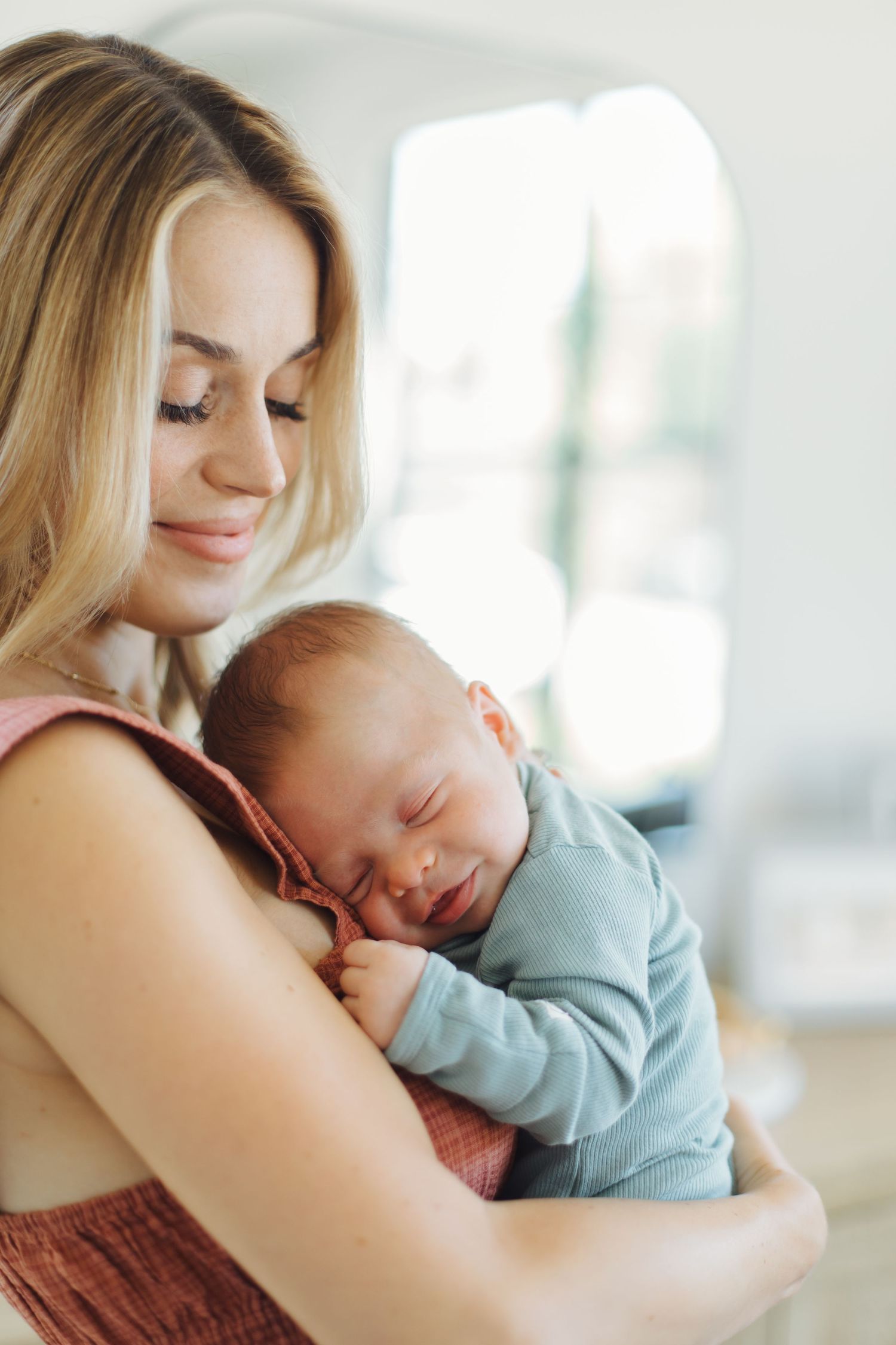 Close-up of loving mother holding sleeping newborn baby in light blue outfit against soft background.