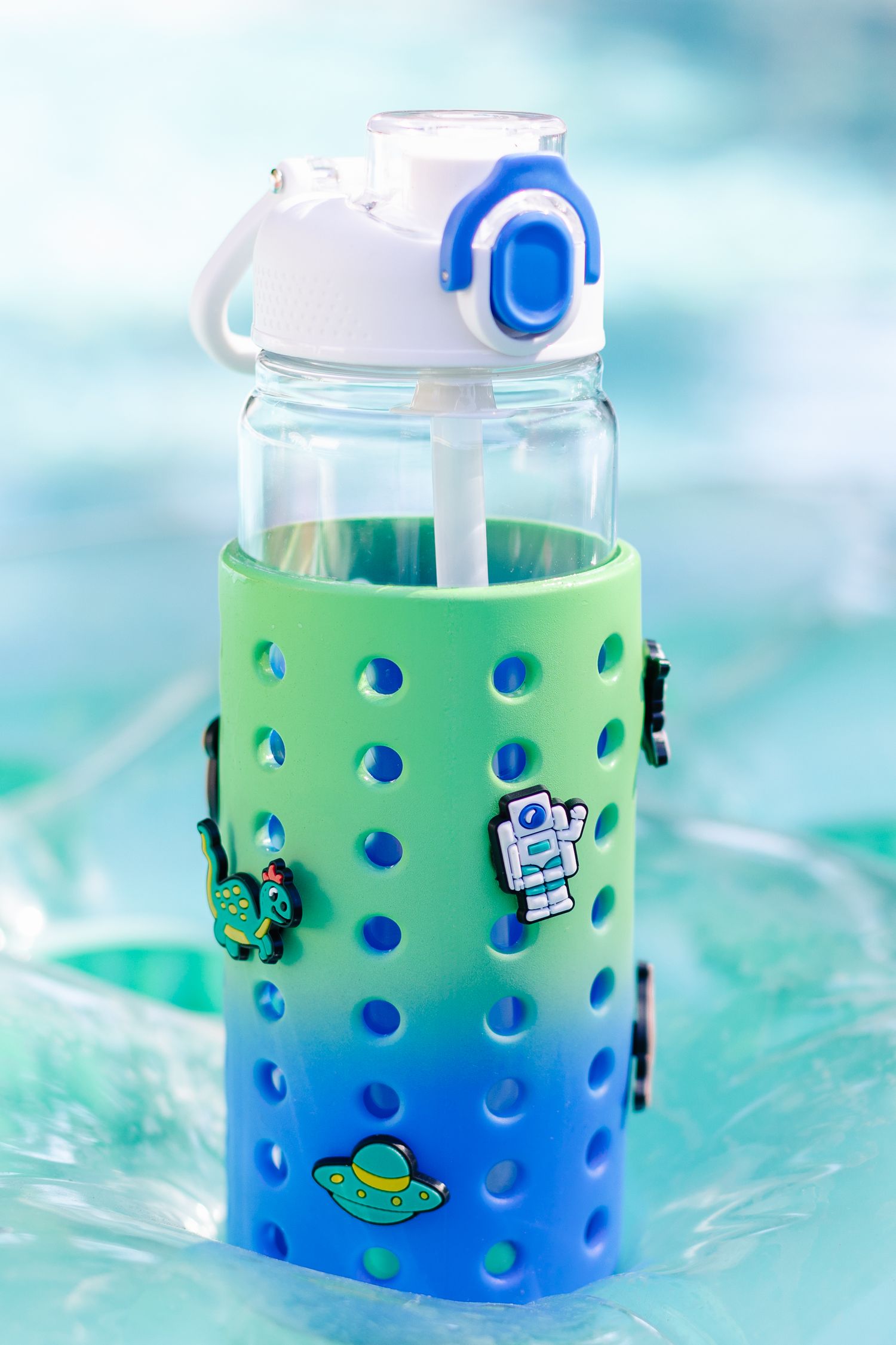 Green and blue ombre silicone sleeve water bottle with astronaut and space themed charms by pool.