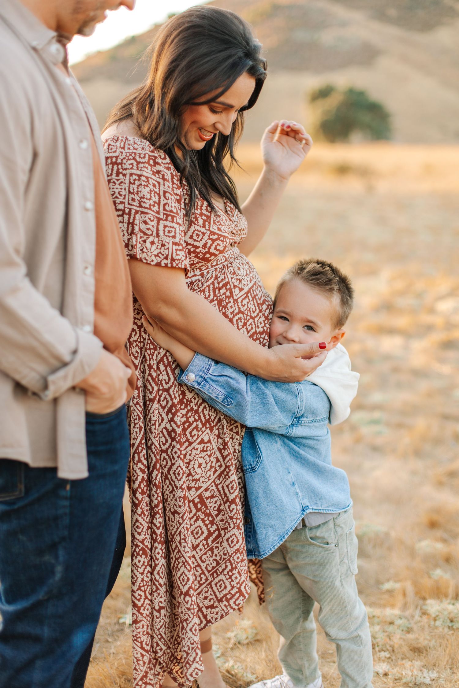 Excited young boy hugging his pregnant mothers baby bump while father stands beside them outdoors.