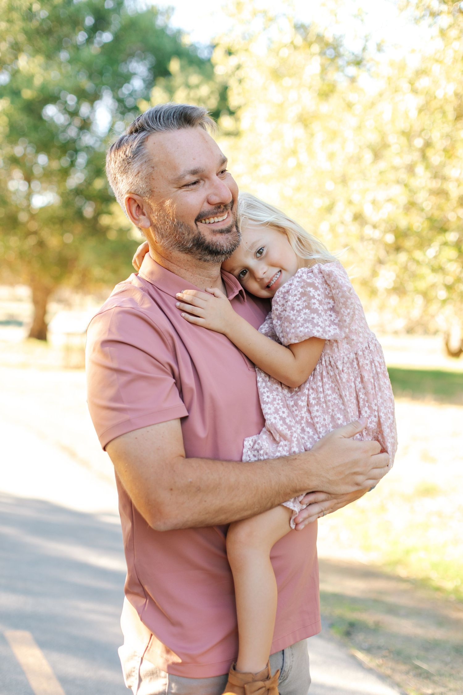 Smiling bearded dad in pink shirt holding his young blonde daughter in a floral dress outdoors.