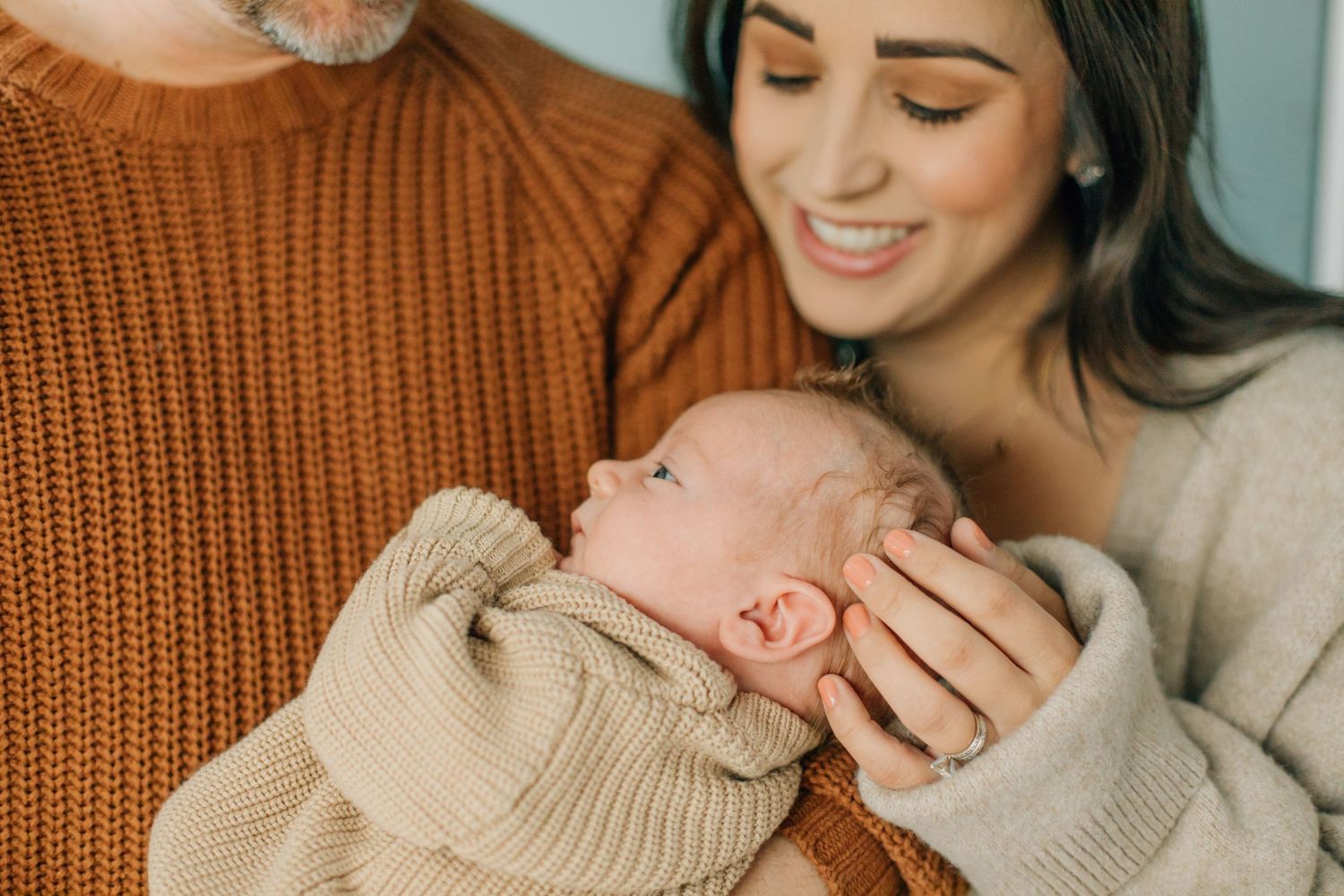 Smiling mother in beige cardigan holding newborn baby with father in rust sweater.
