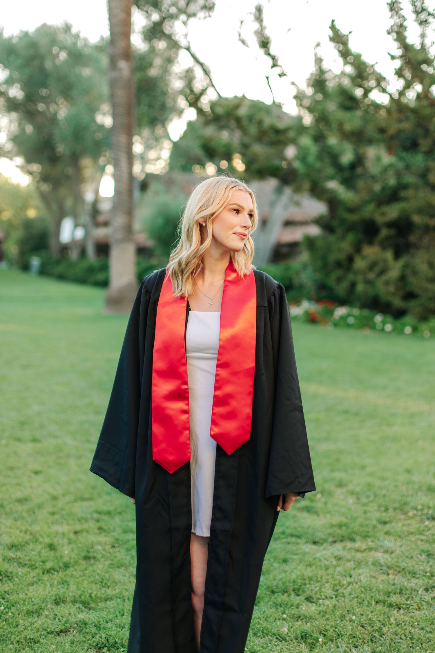Smiling blonde woman in black graduation gown with red stole standing on green grass with trees behind.