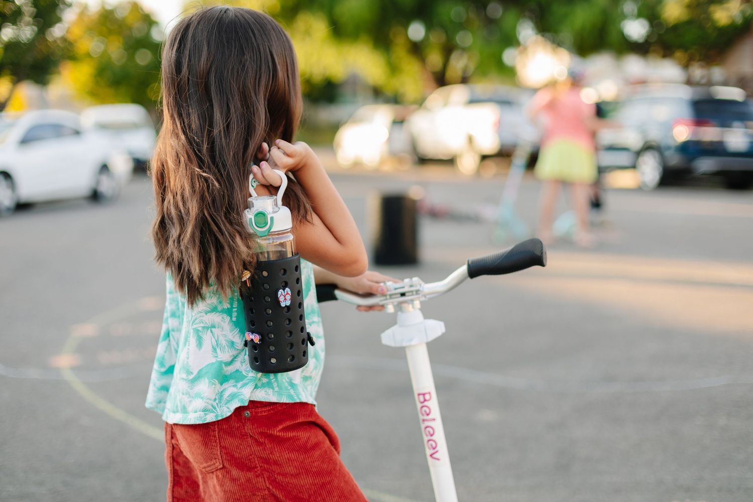Young girl with long hair holding drink bottle standing beside white scooter.