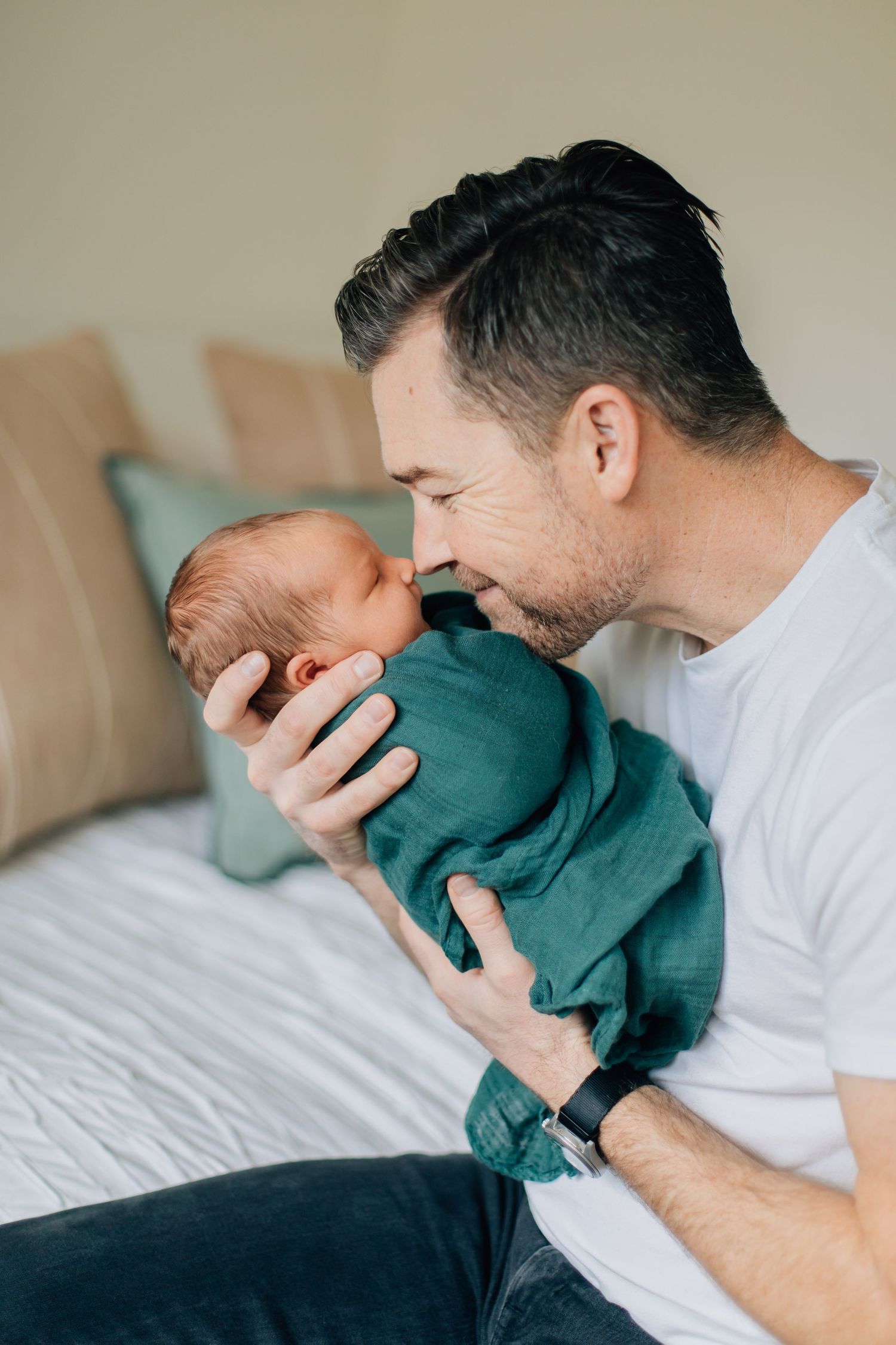 Father tenderly kisses his newborn baby wrapped in a teal swaddle blanket while sitting on a bed.