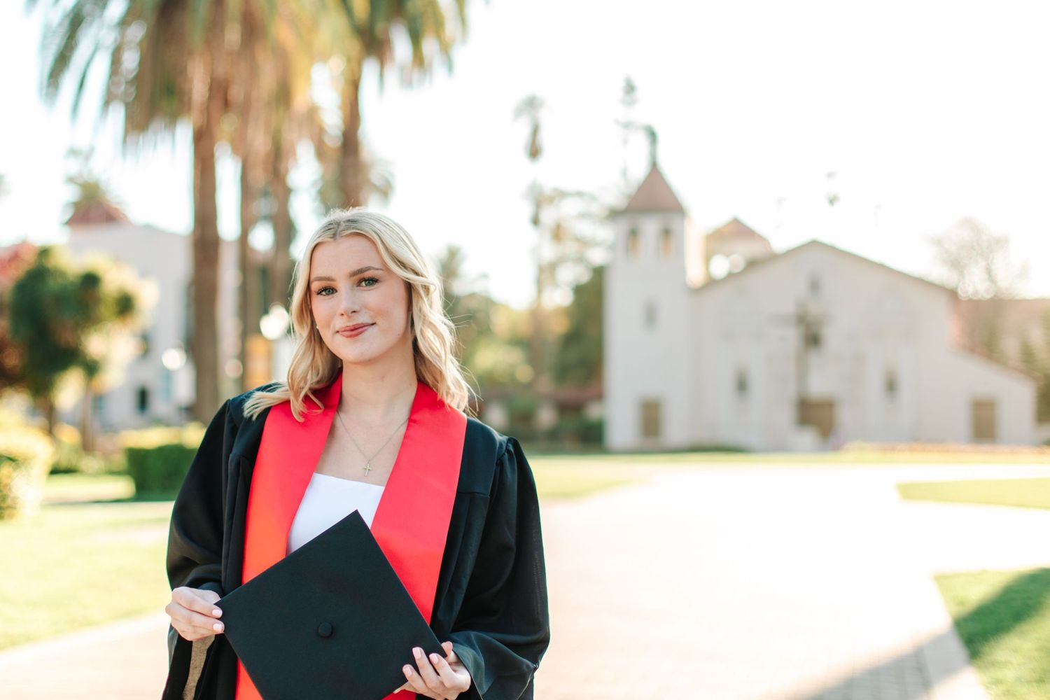 Smiling blonde graduate in black gown and red stole holding diploma near white mission-style church.