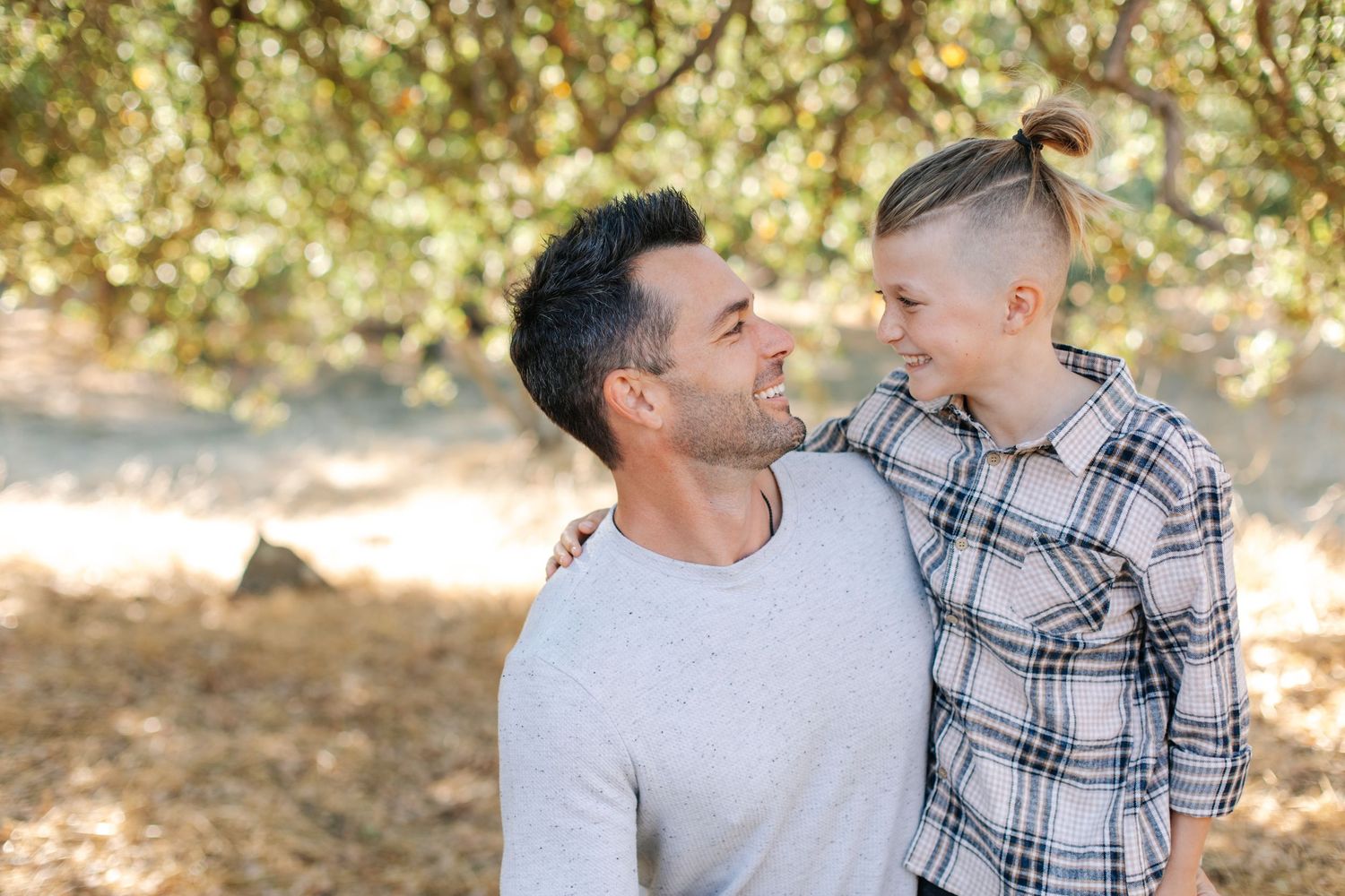 A father and child share a smiling moment together beneath dappled sunlight in a wooded setting.