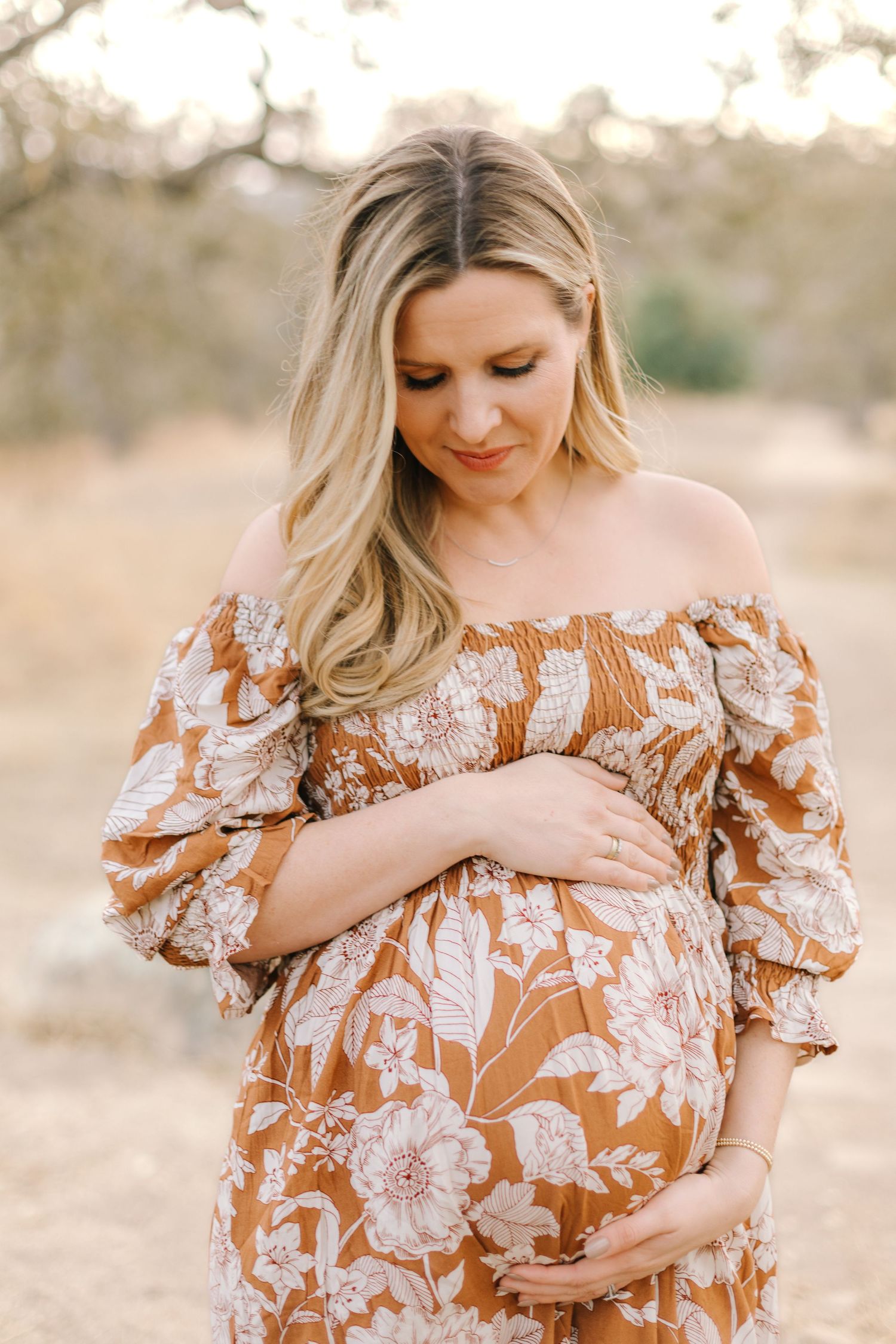 Pregnant woman in rust floral off-shoulder dress gazing down at baby bump in soft natural light.