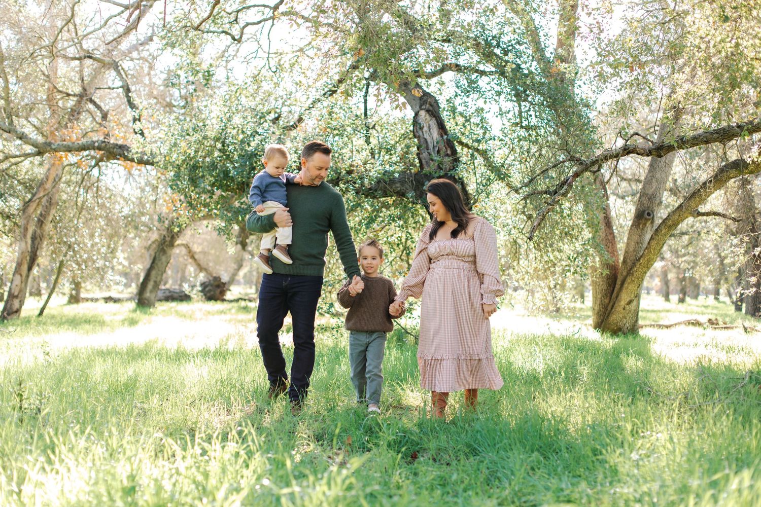 Pregnant mother in pink dress walking with husband and two young children in a green meadow.