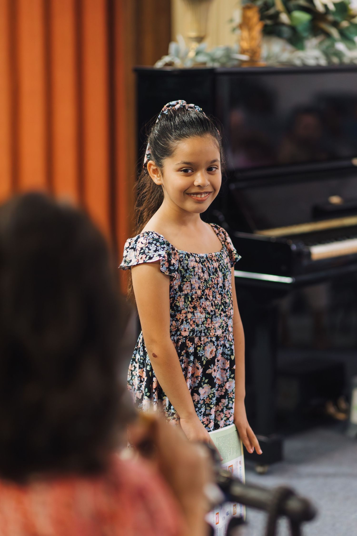 A young girl in a floral dress smiles during a piano recital.