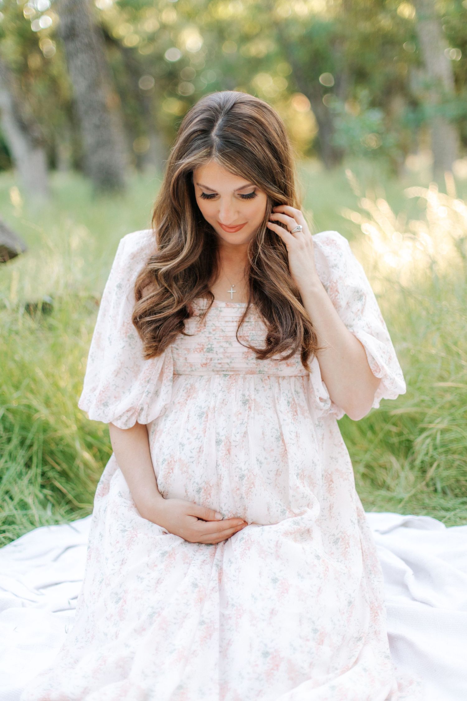 Pregnant woman in floral dress sitting on white blanket touching belly and hair in green meadow.