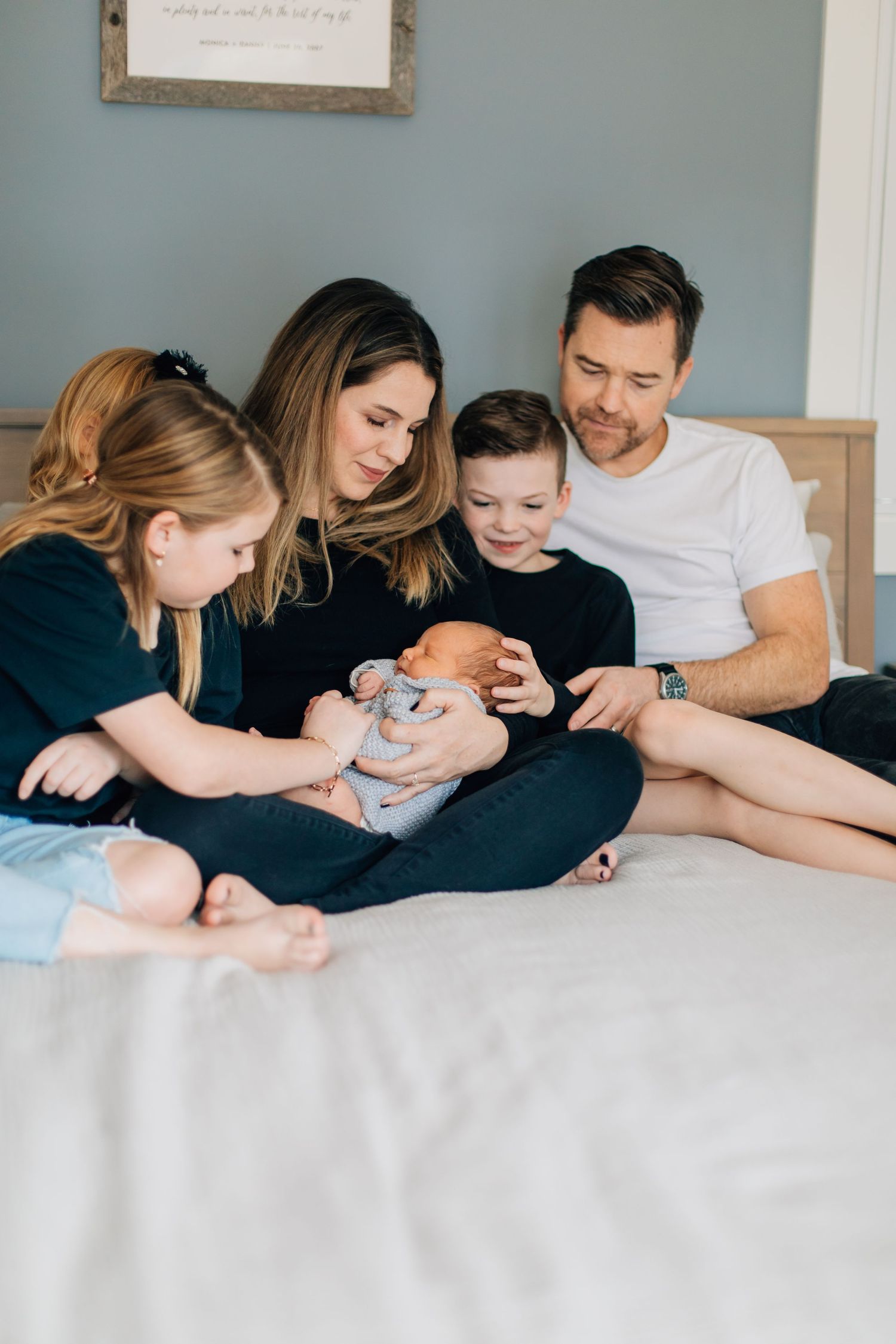 Happy family of four sitting on bed together admiring their newborn baby boy.