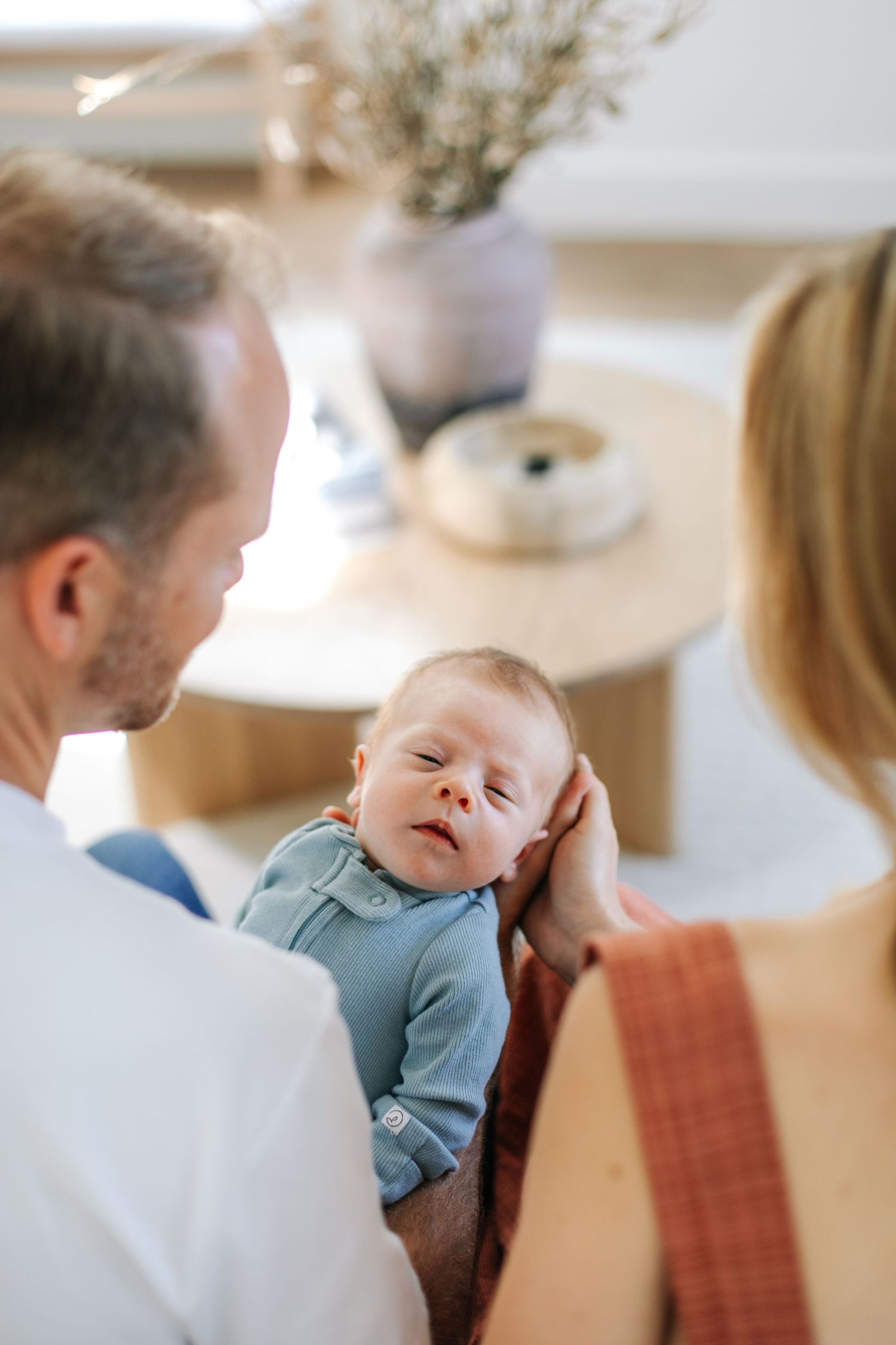 New parents looking down at their alert newborn baby dressed in a light blue outfit at home.