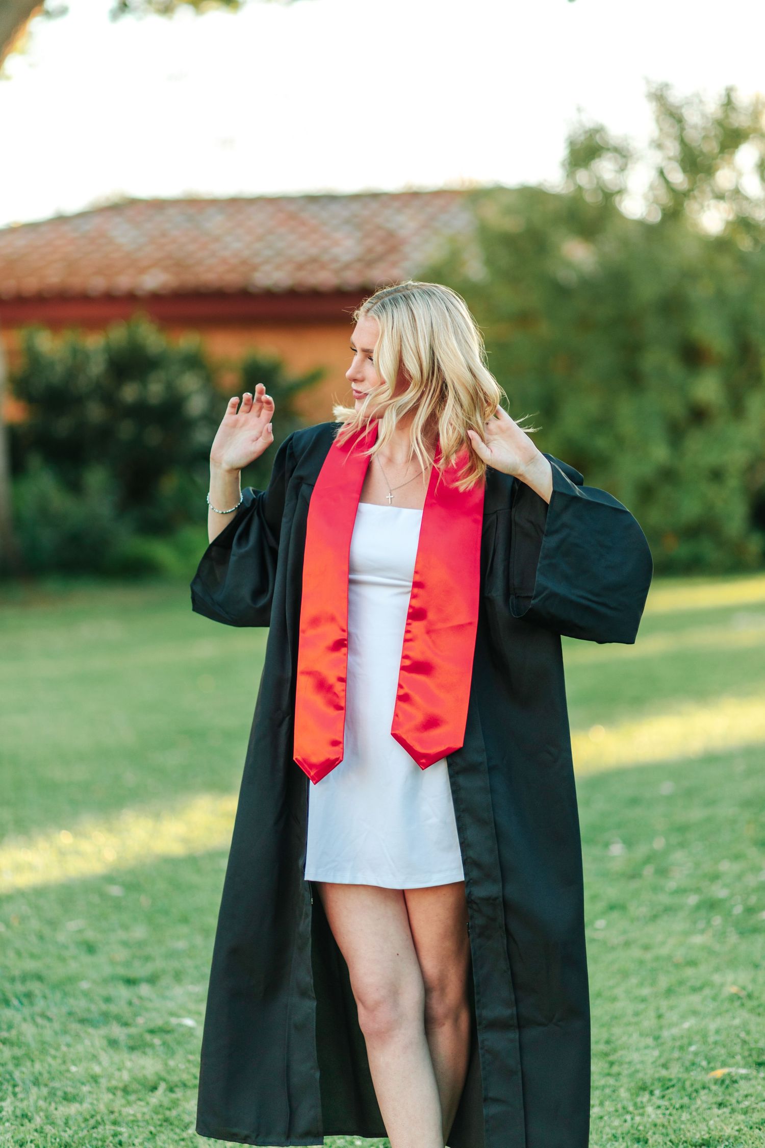 Blonde graduate in open black gown with red stole walking on green lawn near building.