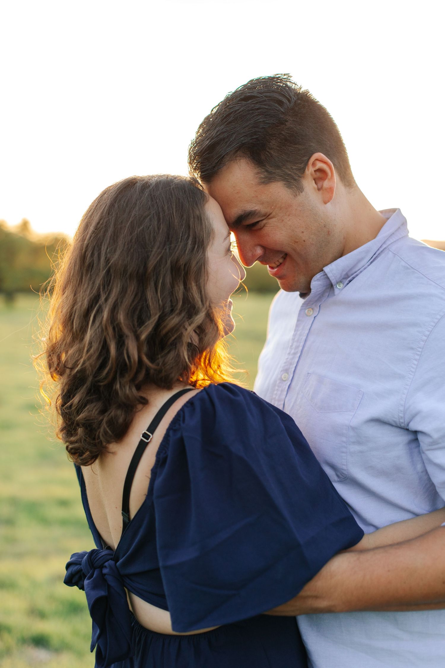 A romantic couple smiles at each other closely, bathed in warm golden sunset light outdoors.