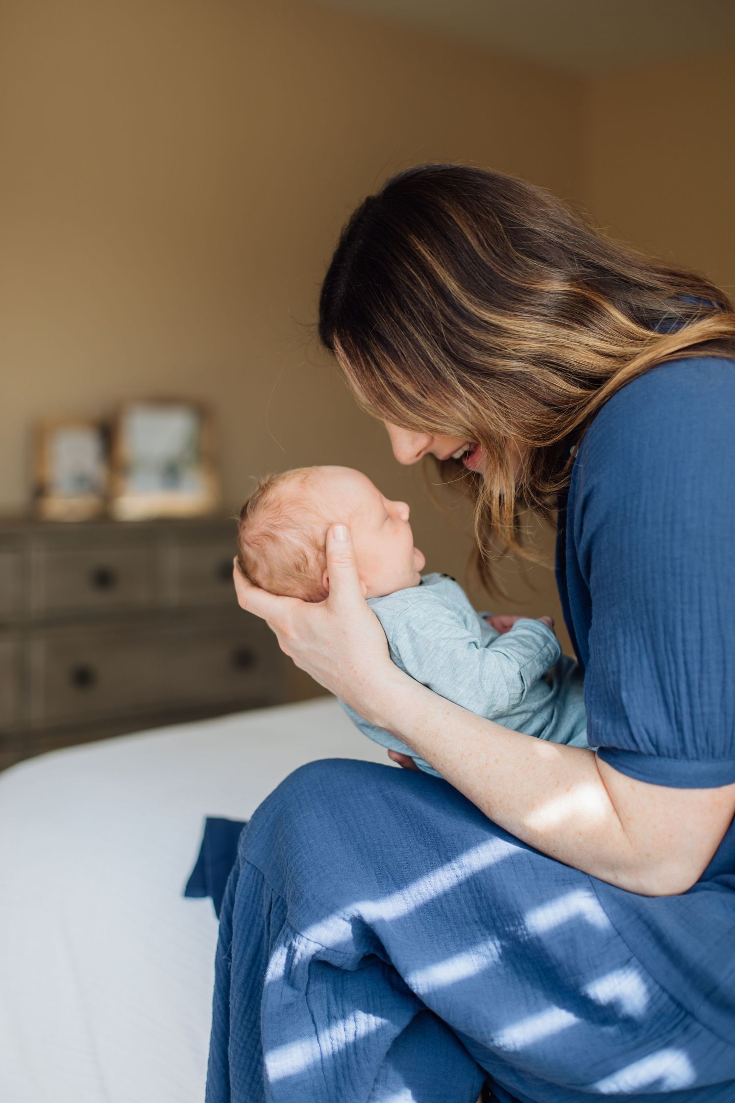 A brunette mother tenderly cradles her newborn baby while seated on a bed in warm sunlight.