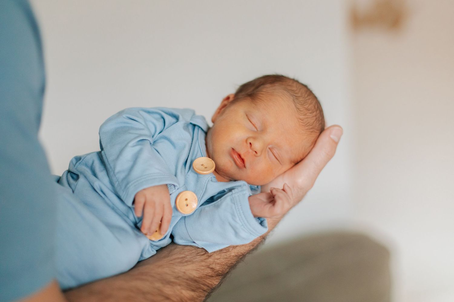 Peaceful newborn baby sleeping in blue button onesie cradled in parent's hands.