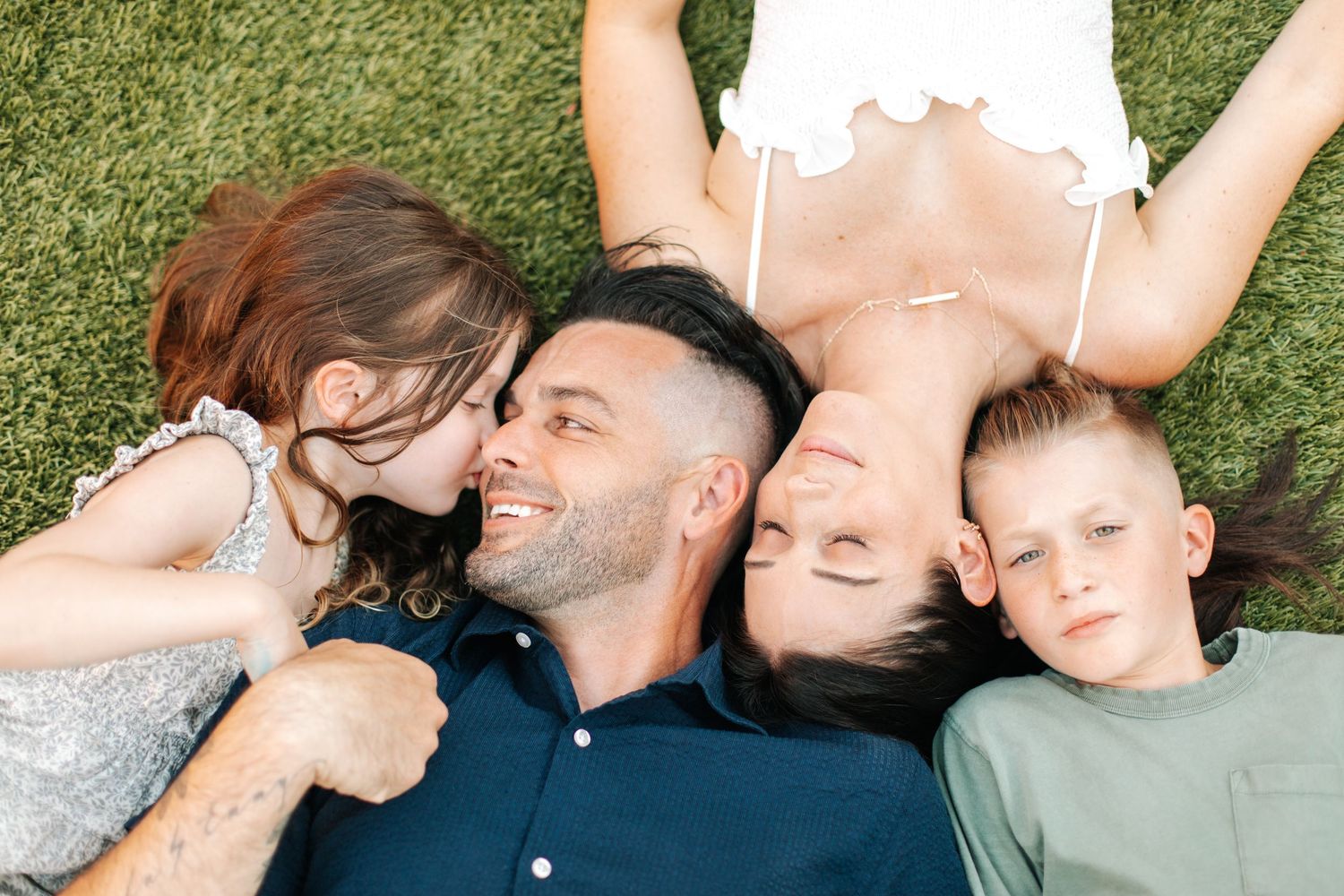 A joyful family of four lies on green grass, smiling and embracing in a top-down view.