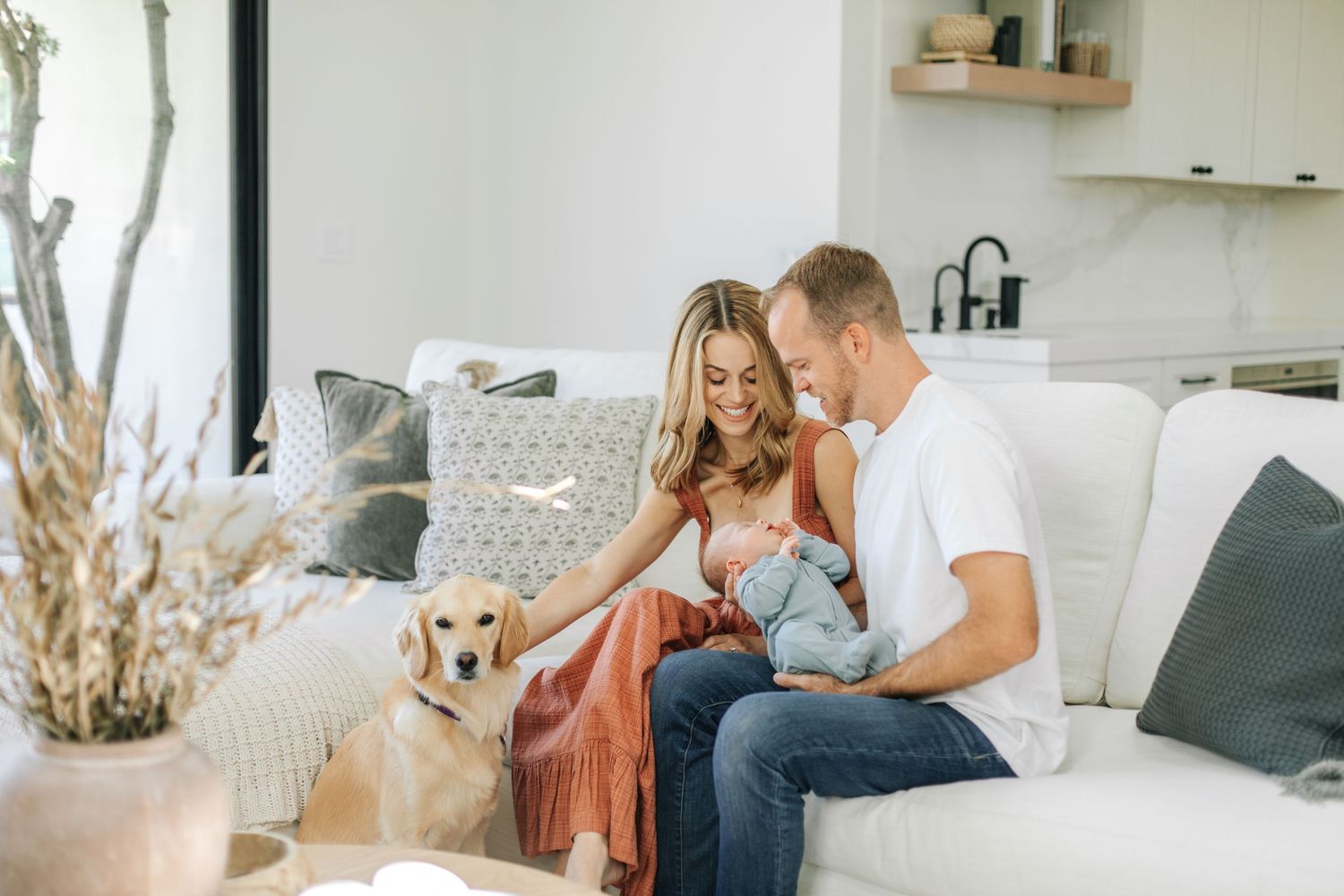 Happy family with newborn baby and golden retriever dog sitting on white sofa in bright modern home.