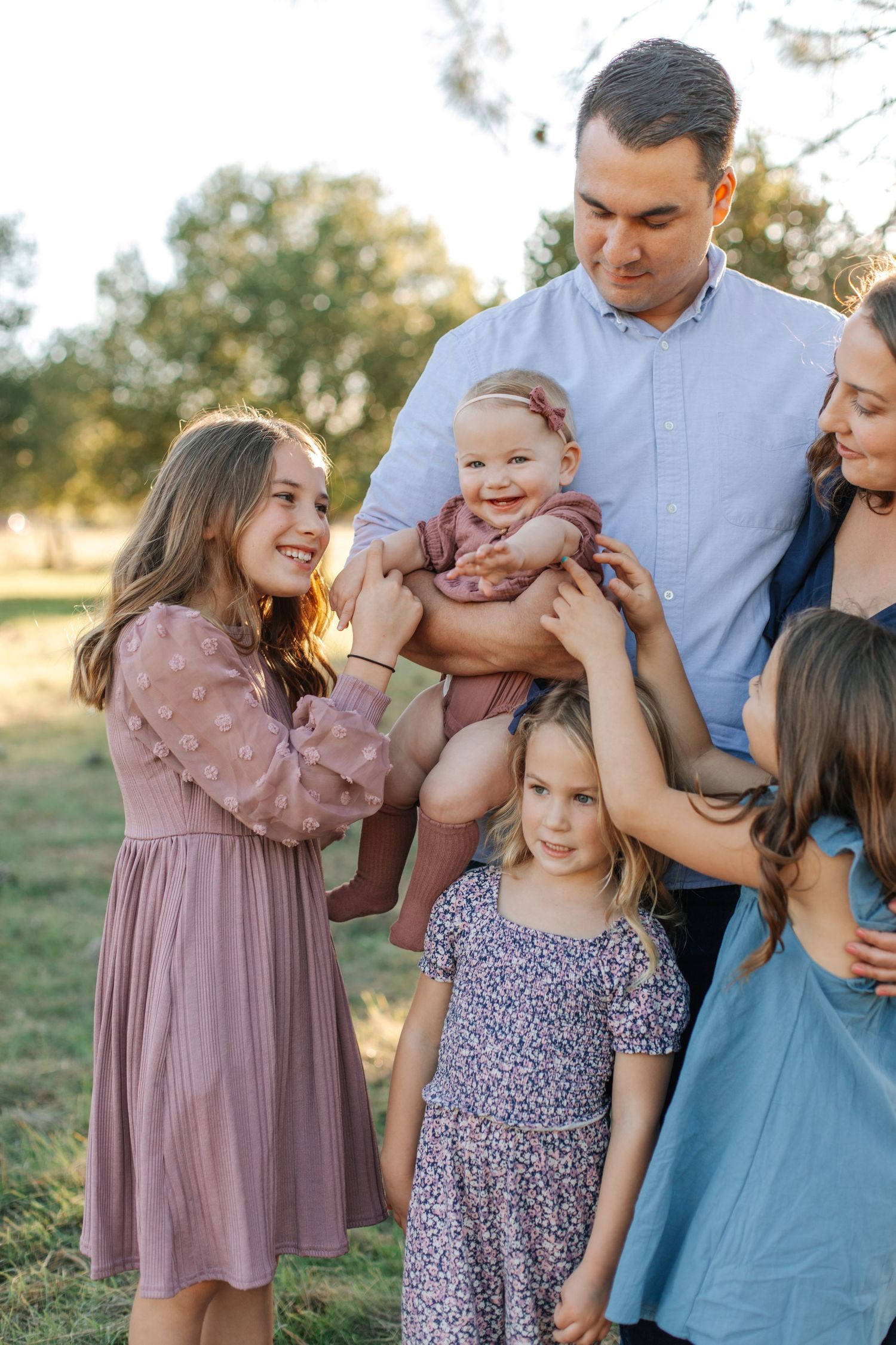 A happy family of six with four daughters, including a baby, poses together in a sunny park.