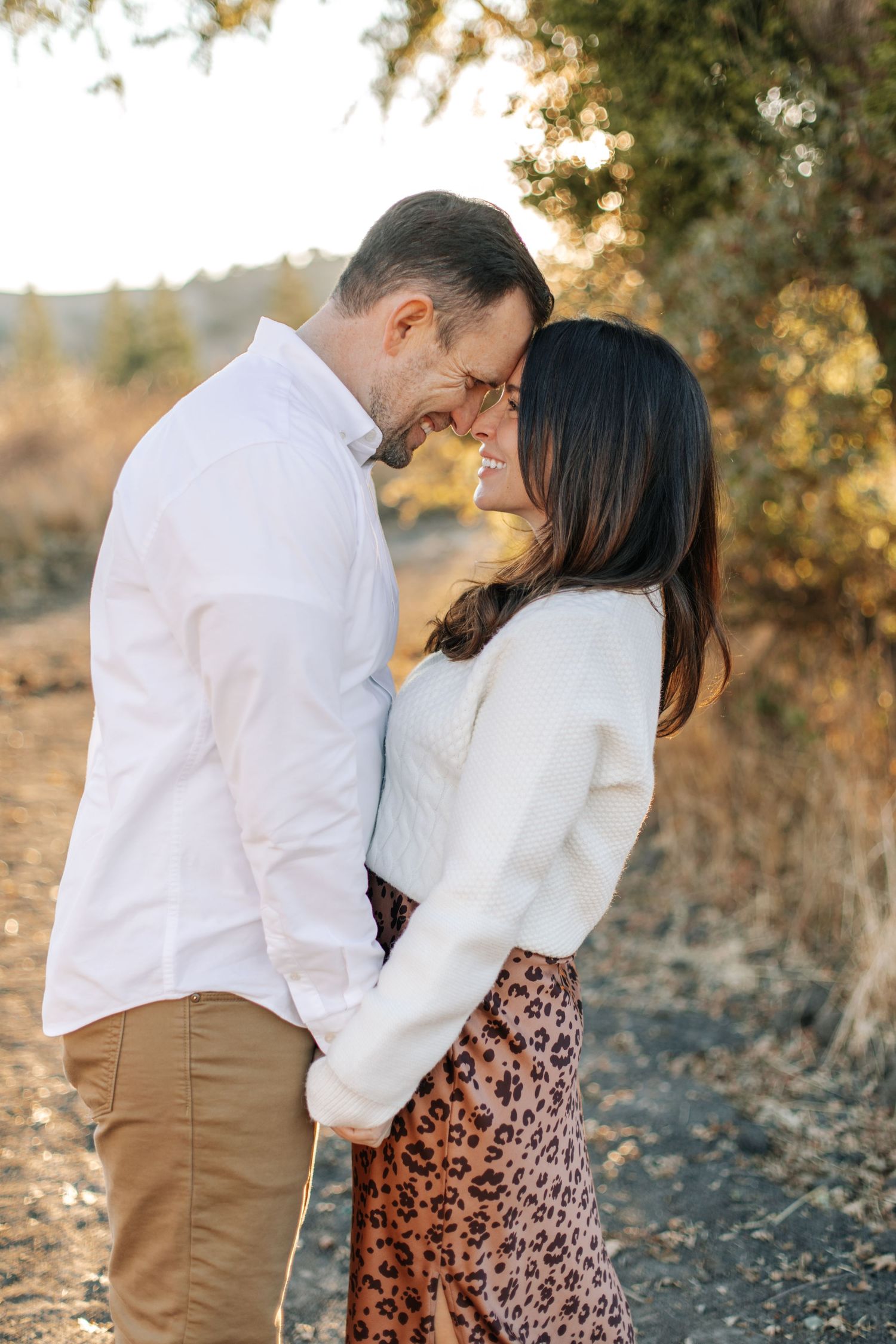 Romantic couple touching foreheads tenderly, woman in white sweater and leopard skirt, golden bokeh background.