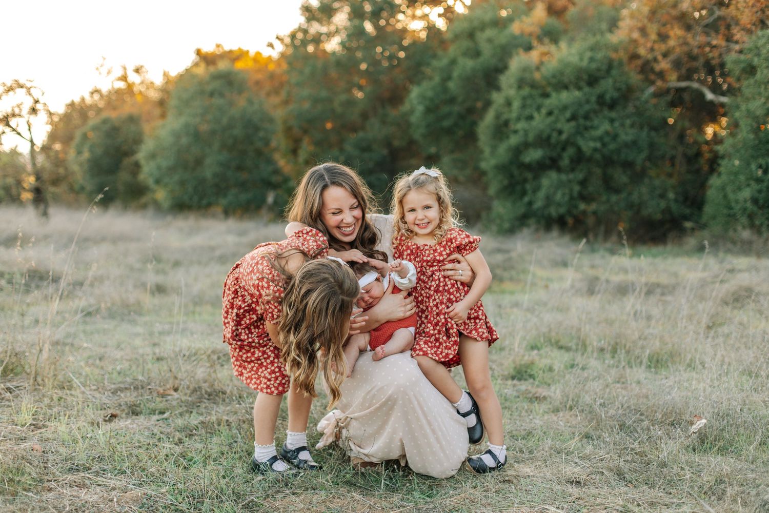 Mother kneeling in grassy field with two young daughters in red dresses cuddling a small dog at sunset.